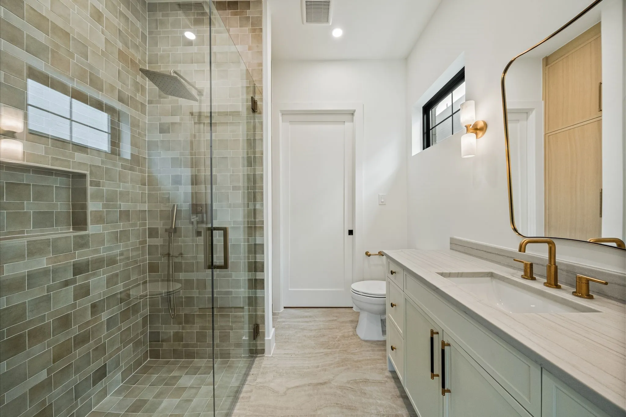Spa-inspired primary bathroom featuring a frameless glass-enclosed shower with subway tile surround, dual vanity with white quartz countertop, brass fixtures, and wood-finish flooring throughout