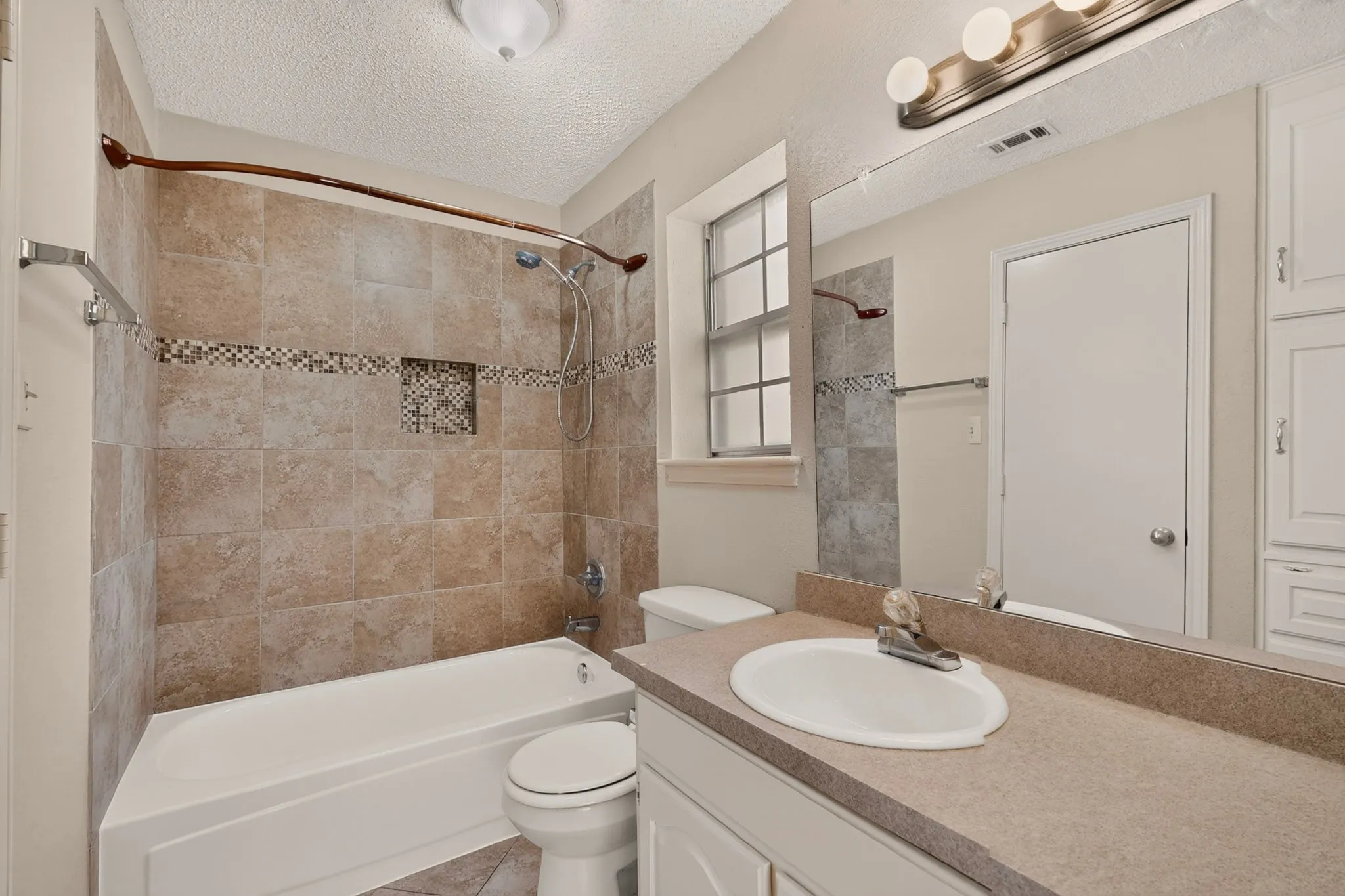 Tile shower surround with mosaic accent, built-in shower niche, single vanity with integrated sink, expansive vanity mirror, and built-in linen cabinetry