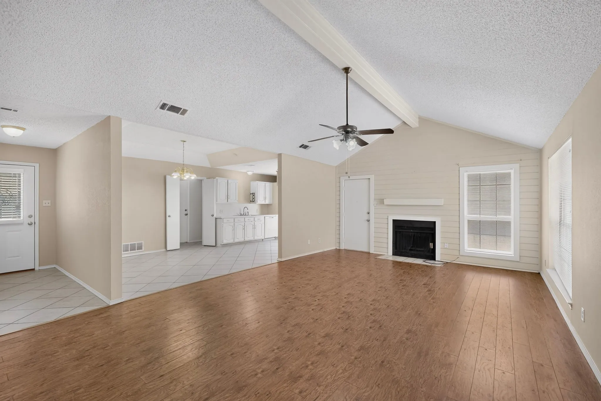 Vaulted ceiling with exposed beam, wood-finish flooring, fireplace with mantel, ceiling fan, and white kitchen cabinetry with tile flooring