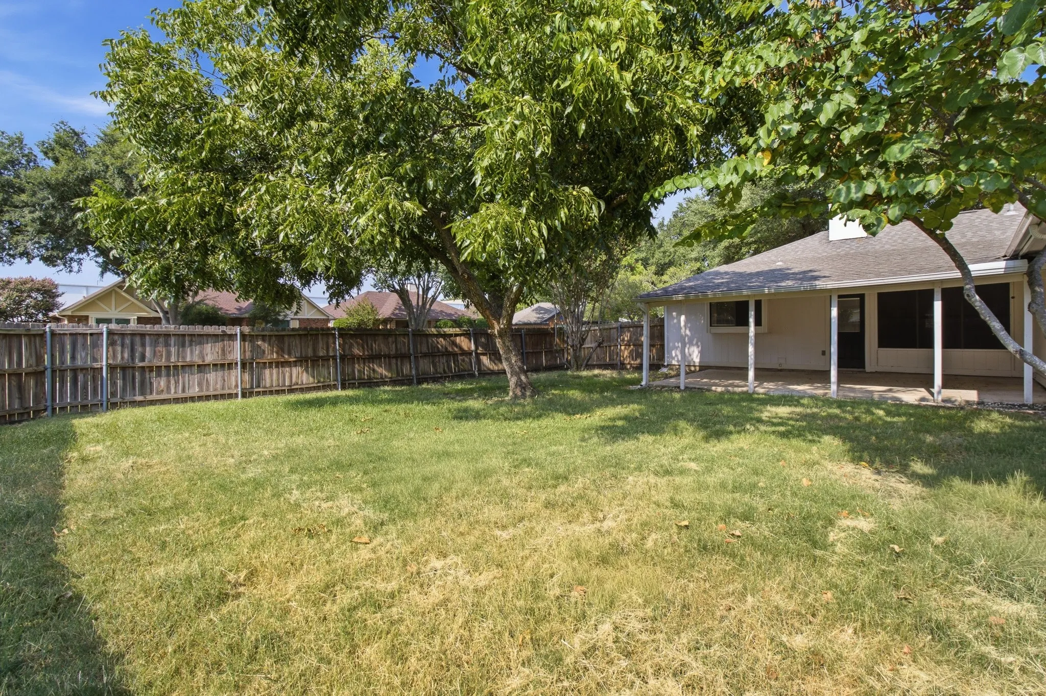 Expansive backyard featuring a wood privacy fence, mature trees, and a covered concrete patio