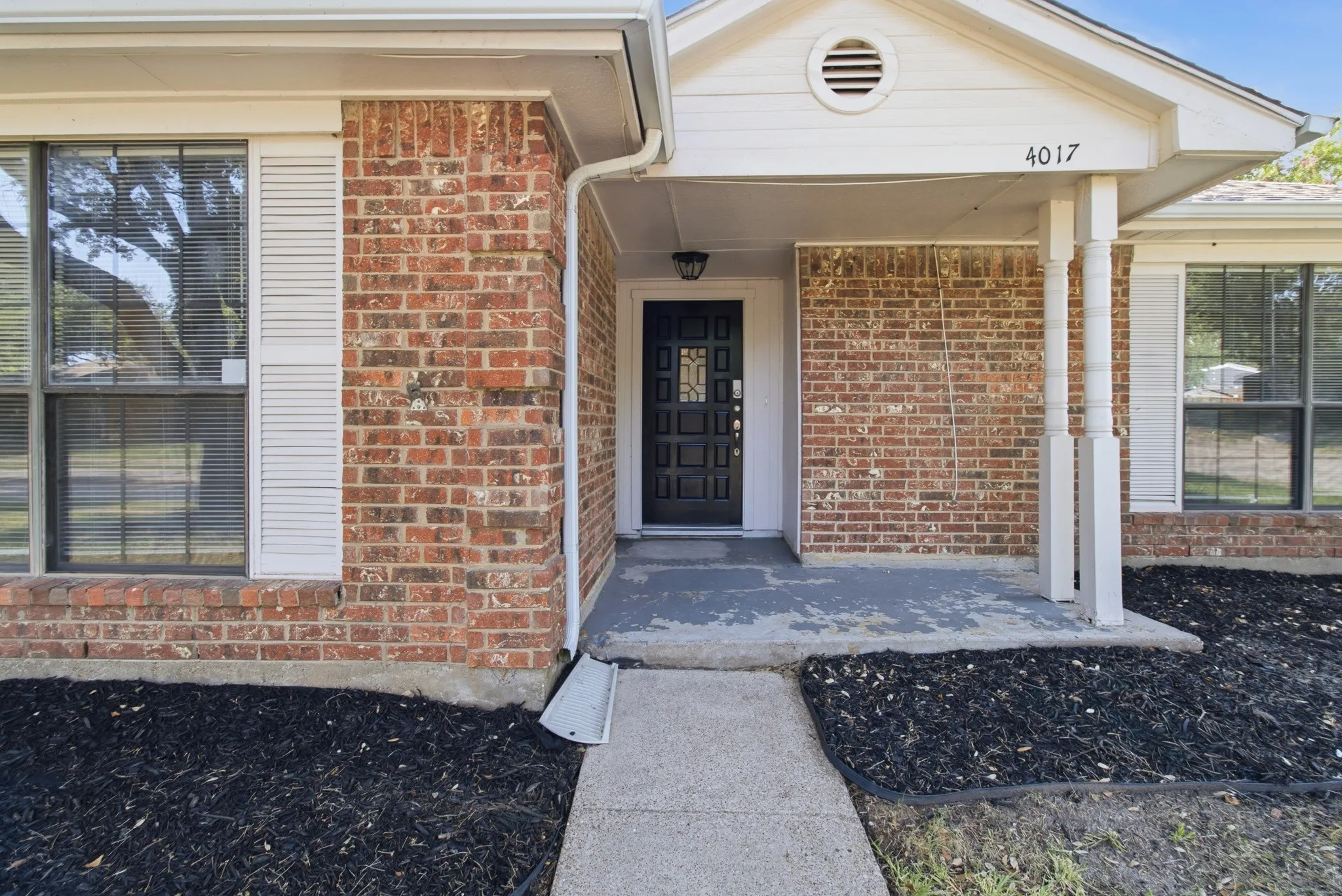 Brick facade home with a covered front entry featuring a black multi-pane door, white window shutters, and a concrete walkway