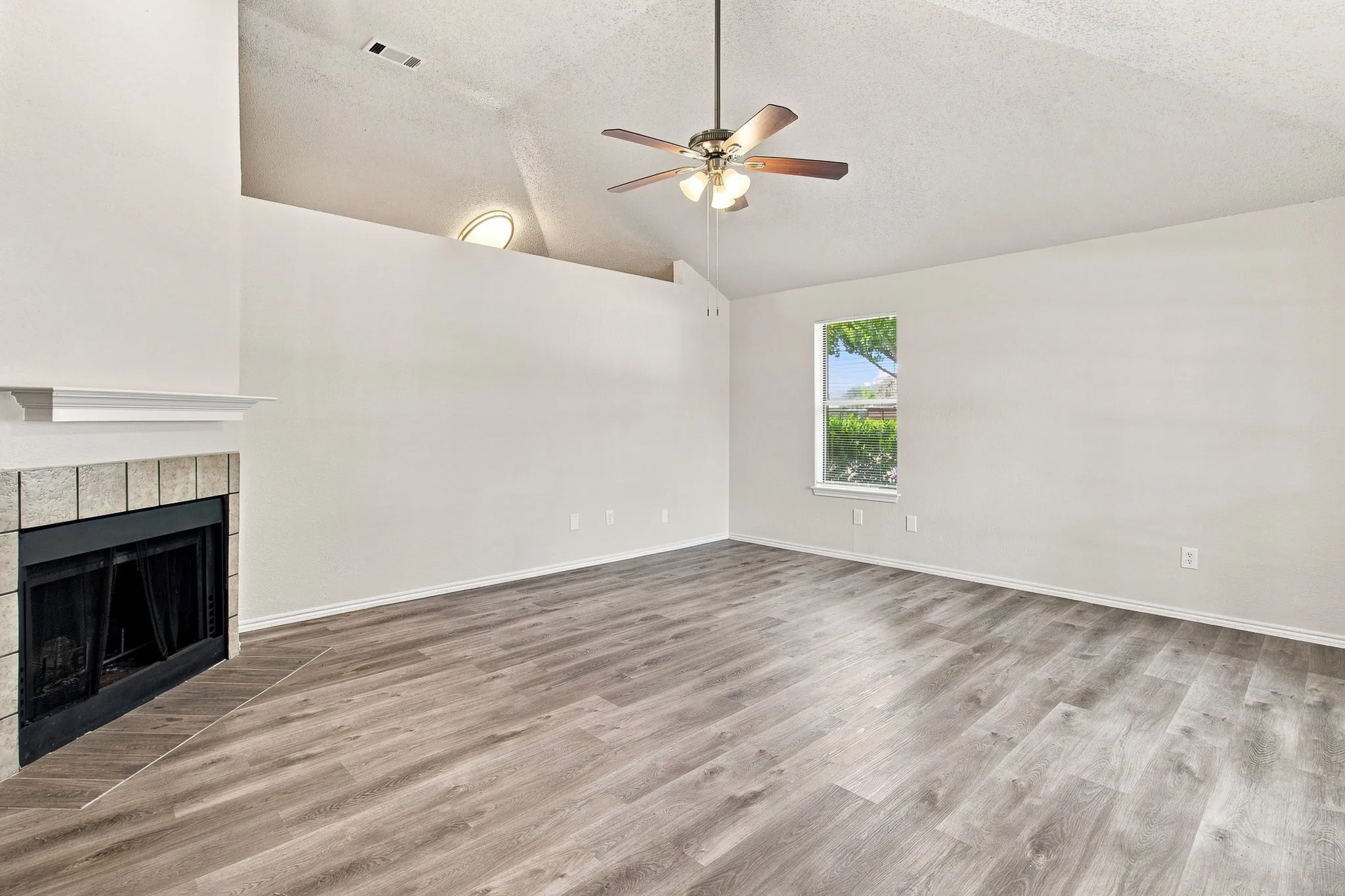 Spacious living area featuring wood-finish flooring, a tiled fireplace with mantel, a vaulted ceiling with a ceiling fan, and a prominent window
