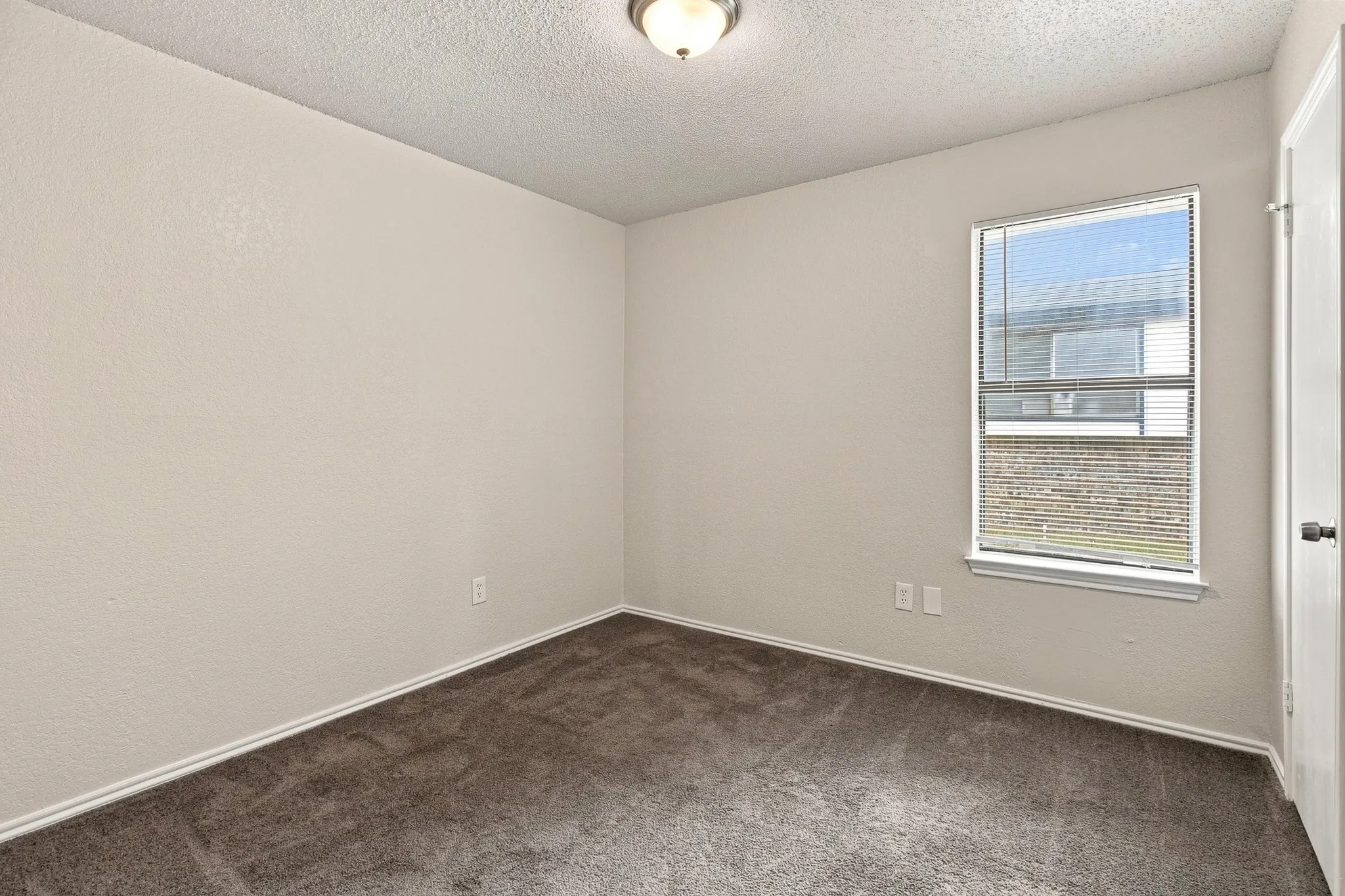 Carpeted interior room featuring light-colored walls, a single window with horizontal blinds, and a flush-mount ceiling light fixture