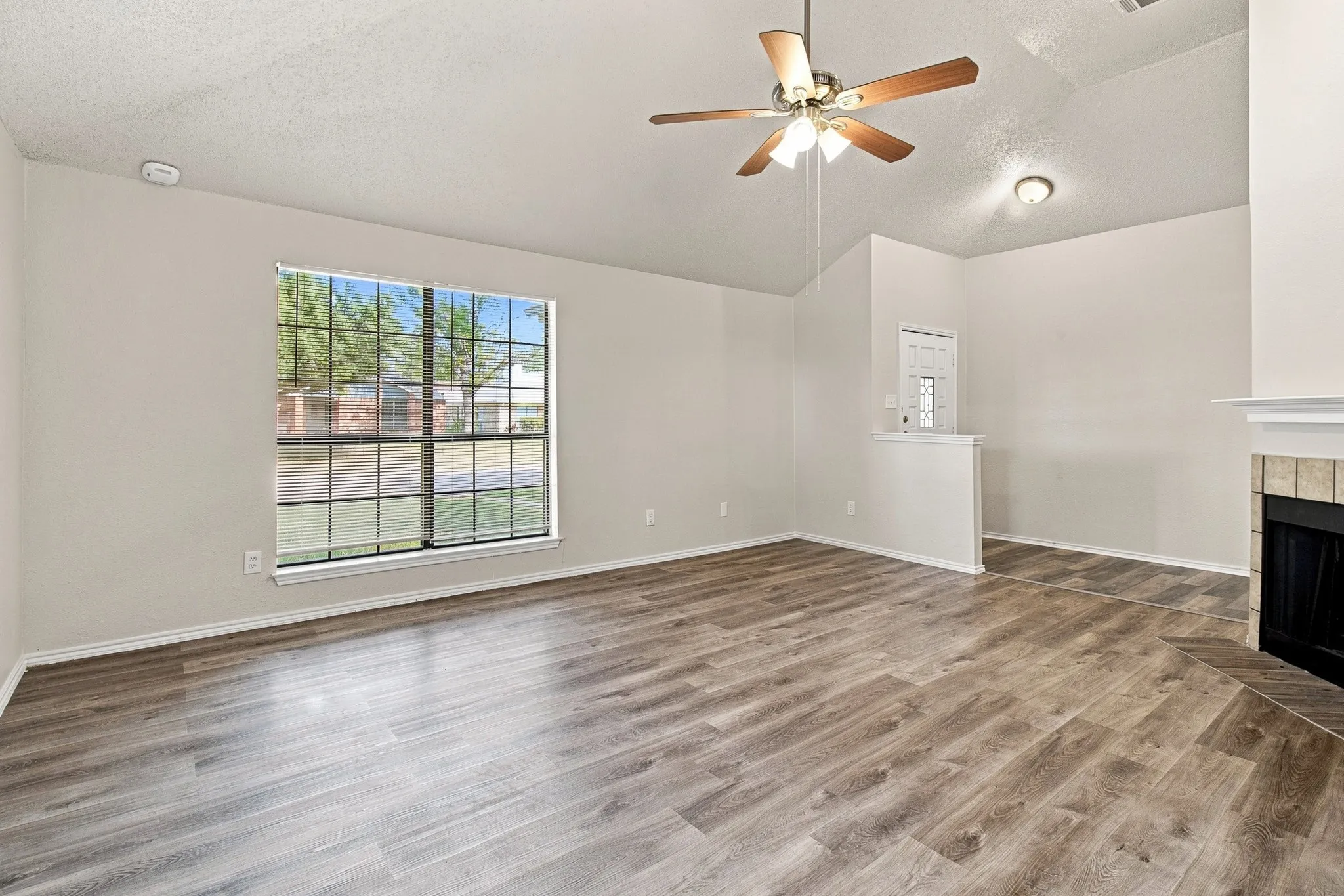 Spacious interior featuring wood-finish flooring, a large window with grid pattern, a ceiling fan with integrated lighting, a fireplace with a stone-tile surround, and vaulted ceilings