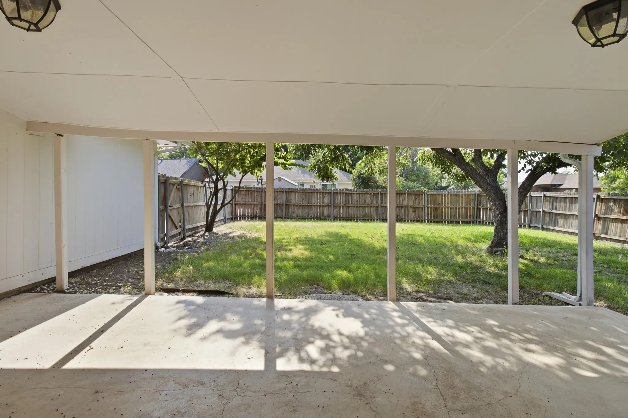 Covered patio with concrete flooring, supported by white posts, opening to a fenced backyard with mature trees and a lawn