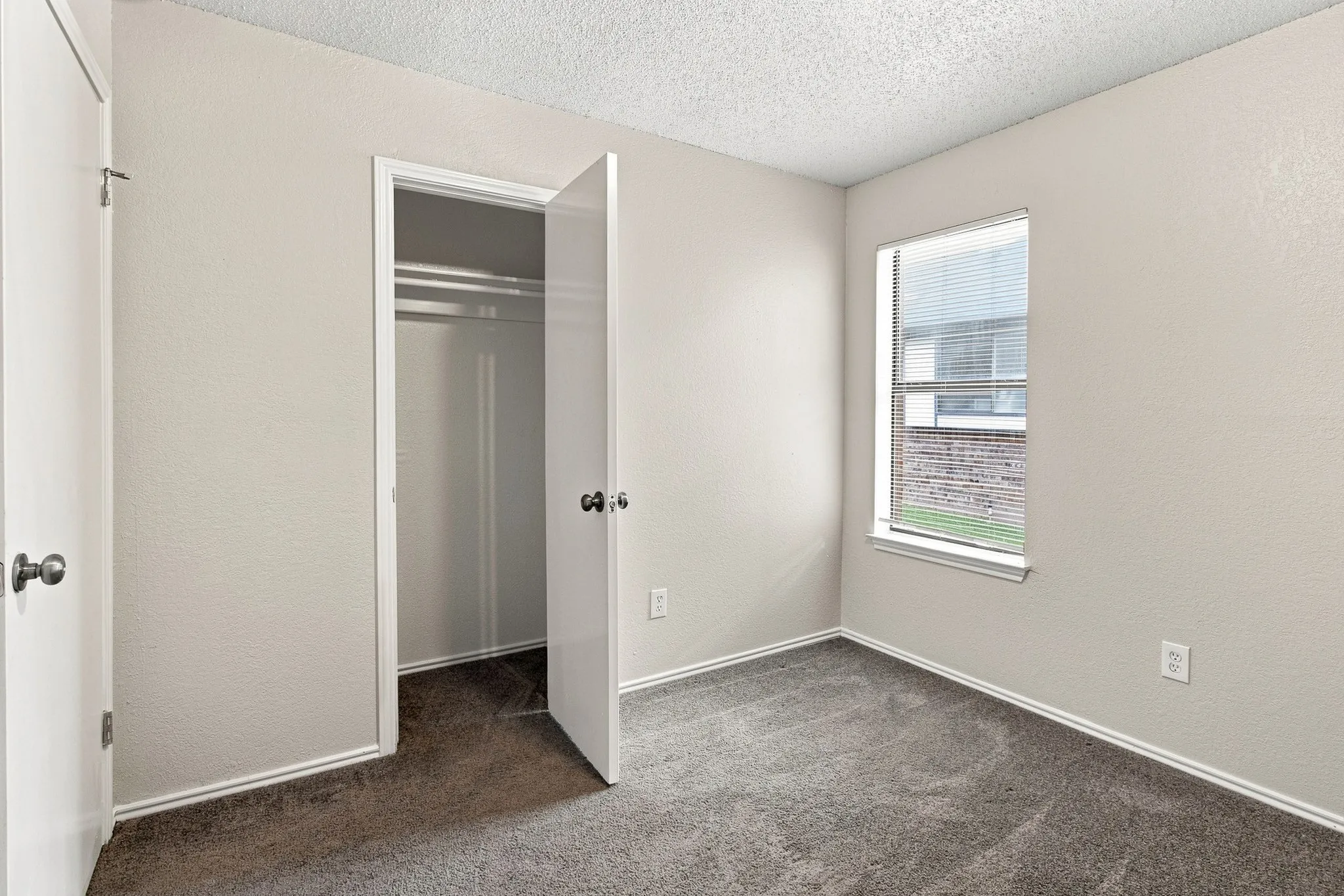 Carpeted room featuring a window with blinds, light-toned walls, white trim, and a closet with a wood-finish floor