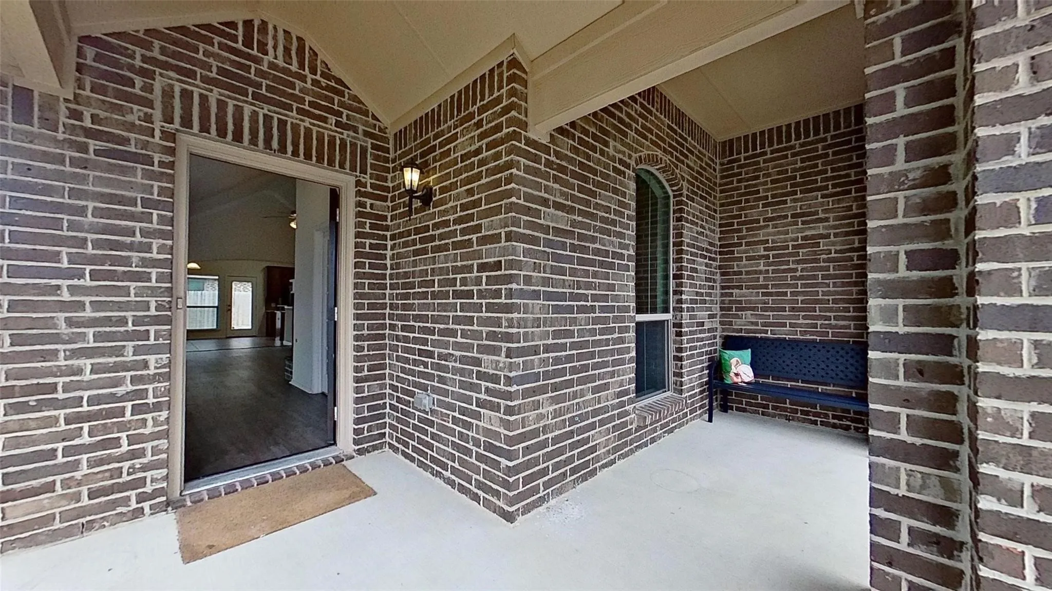 Inviting covered porch featuring dark brickwork, a concrete floor, an arched window with blinds, and a mounted exterior light fixture