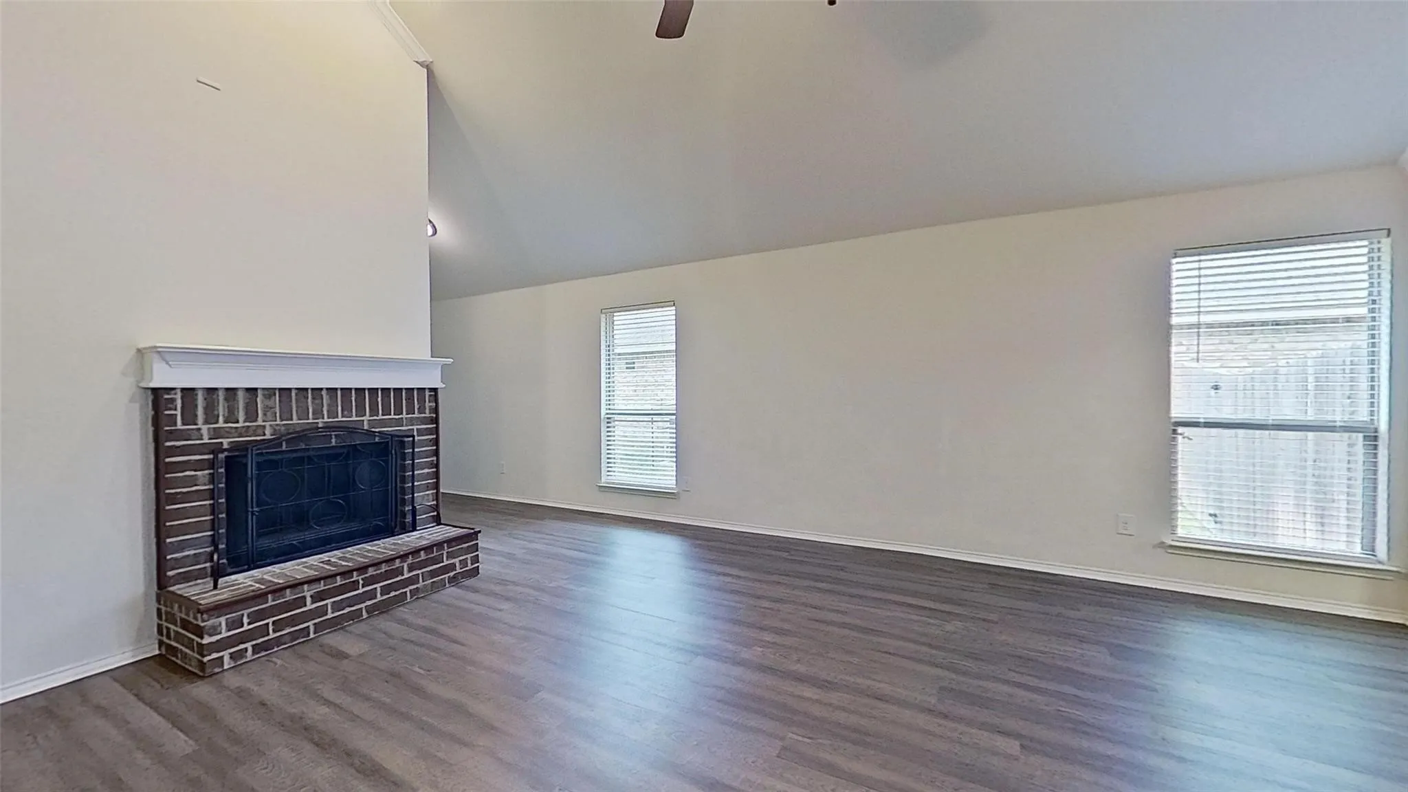 Living area with a vaulted ceiling, wood-finish flooring, a brick fireplace with a white mantel, and two windows with blinds