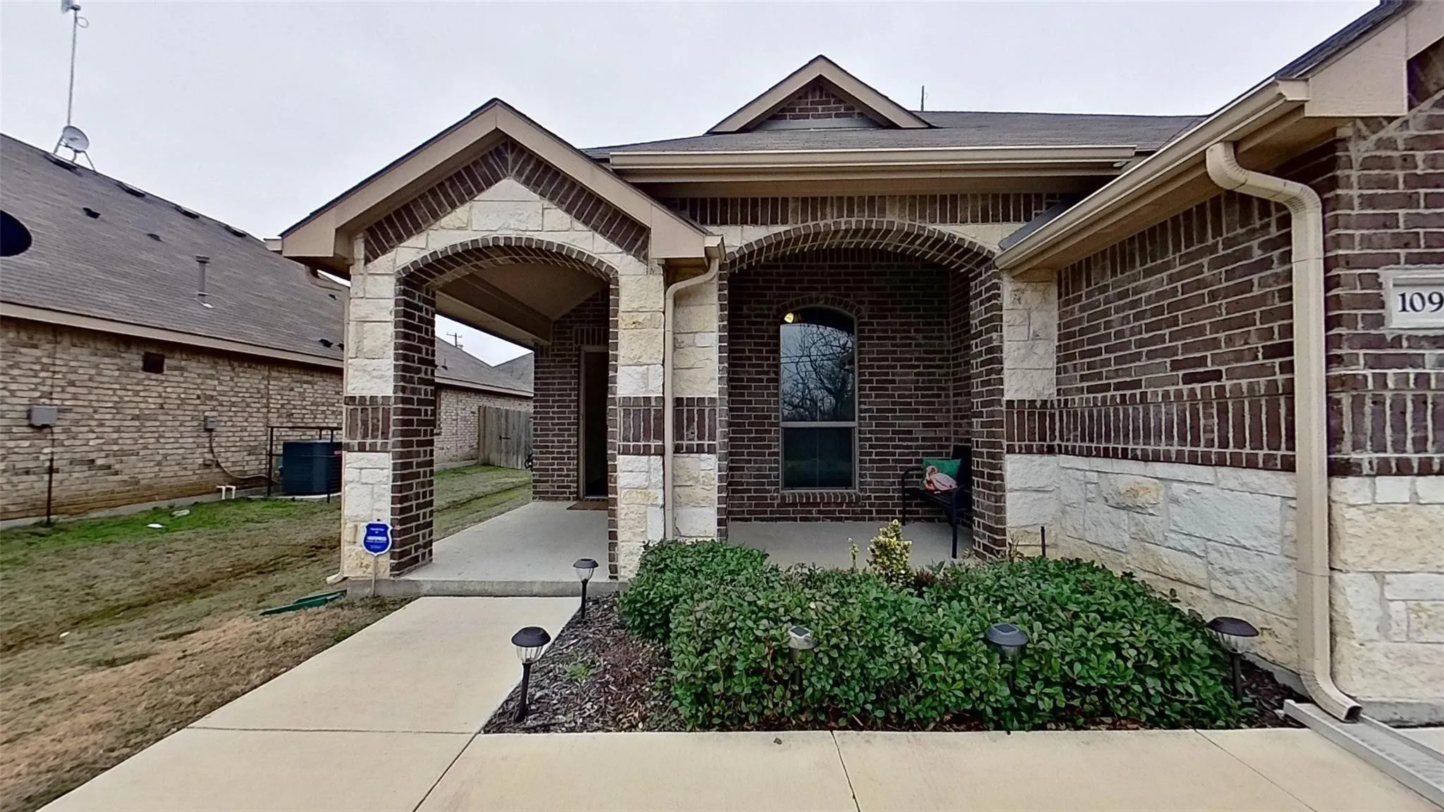 Welcoming front entry showcasing a brick and stone exterior with arched architectural detailing, a covered porch with decorative railings, and professionally landscaped foundation plantings