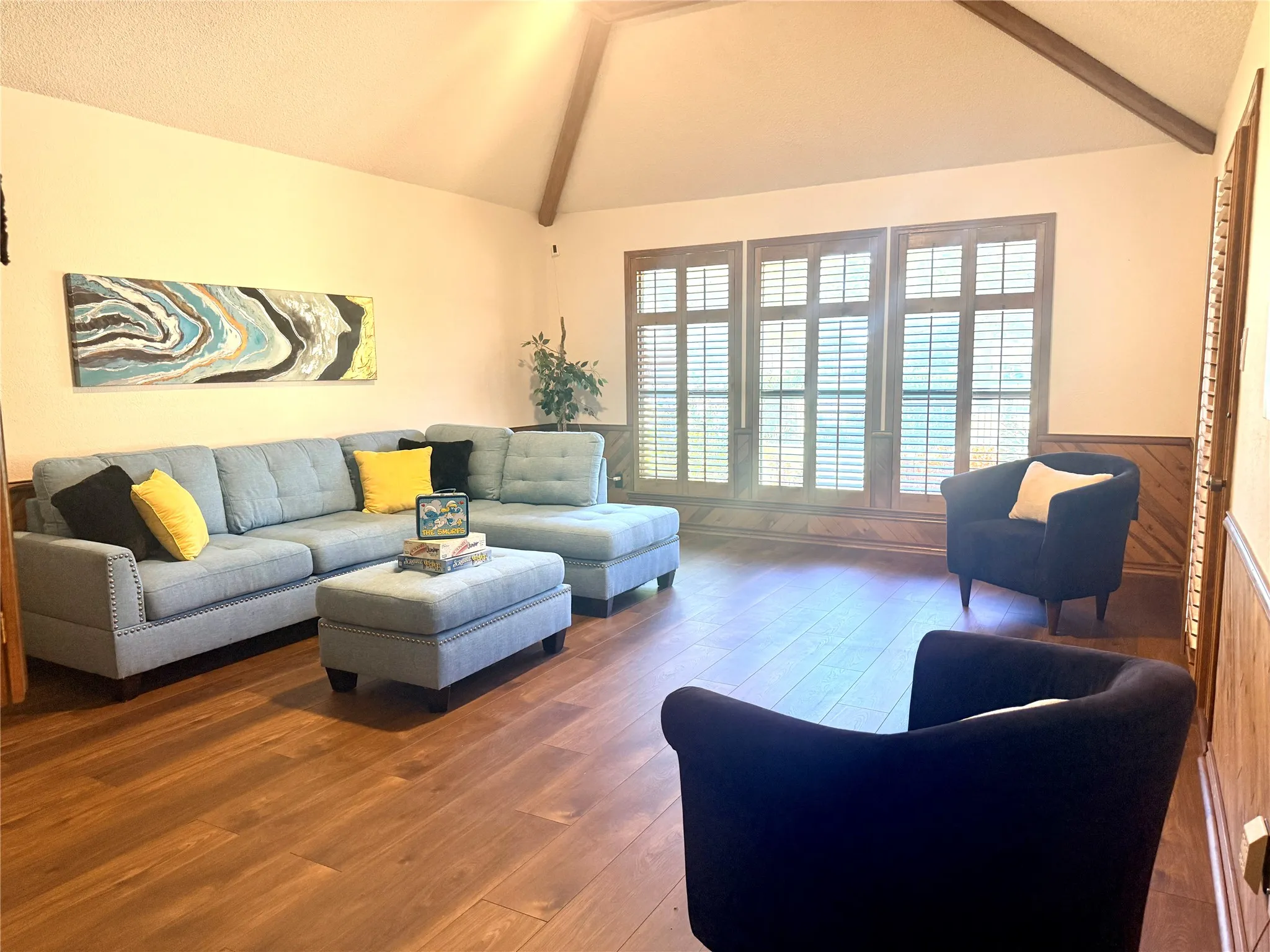 Vaulted ceiling with exposed wood beams, wood-finish flooring, wood paneling, and multiple windows with plantation shutters