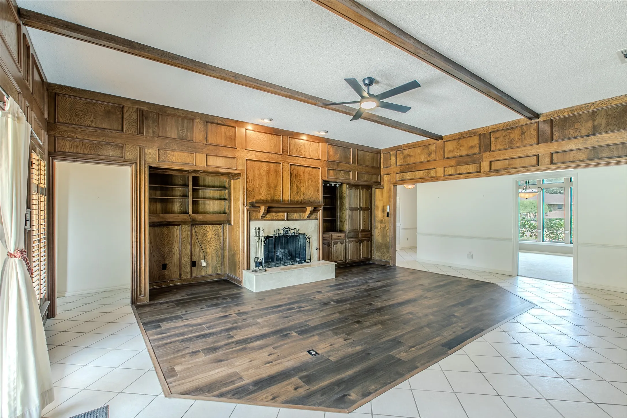Living area featuring wood paneling, exposed ceiling beams, a stone fireplace with an ornate screen, built-in shelving, and a modern ceiling fan