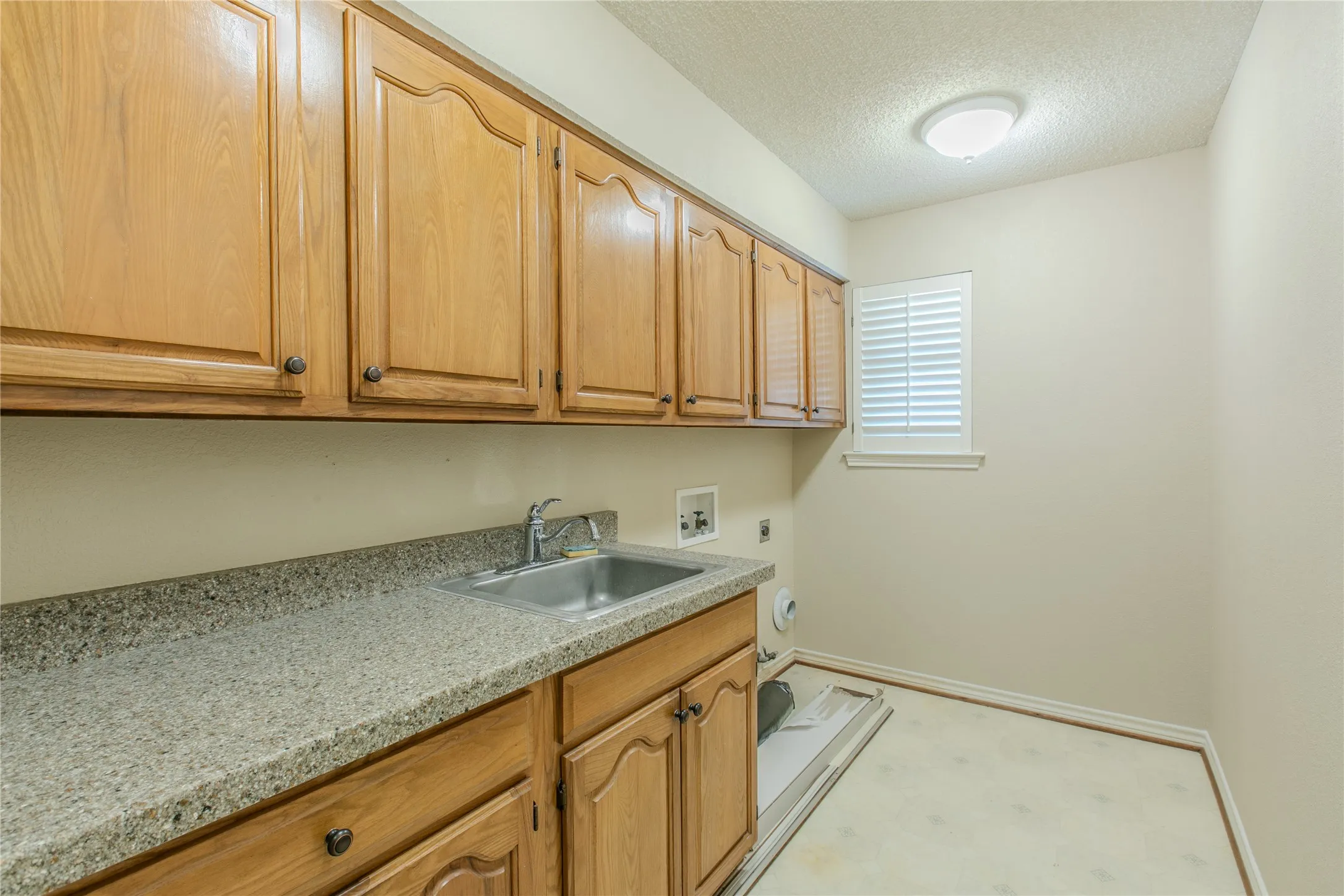 Utility room featuring extensive wood-finish cabinetry, a stone-finish countertop with an integrated stainless steel sink, and a window with plantation shutters