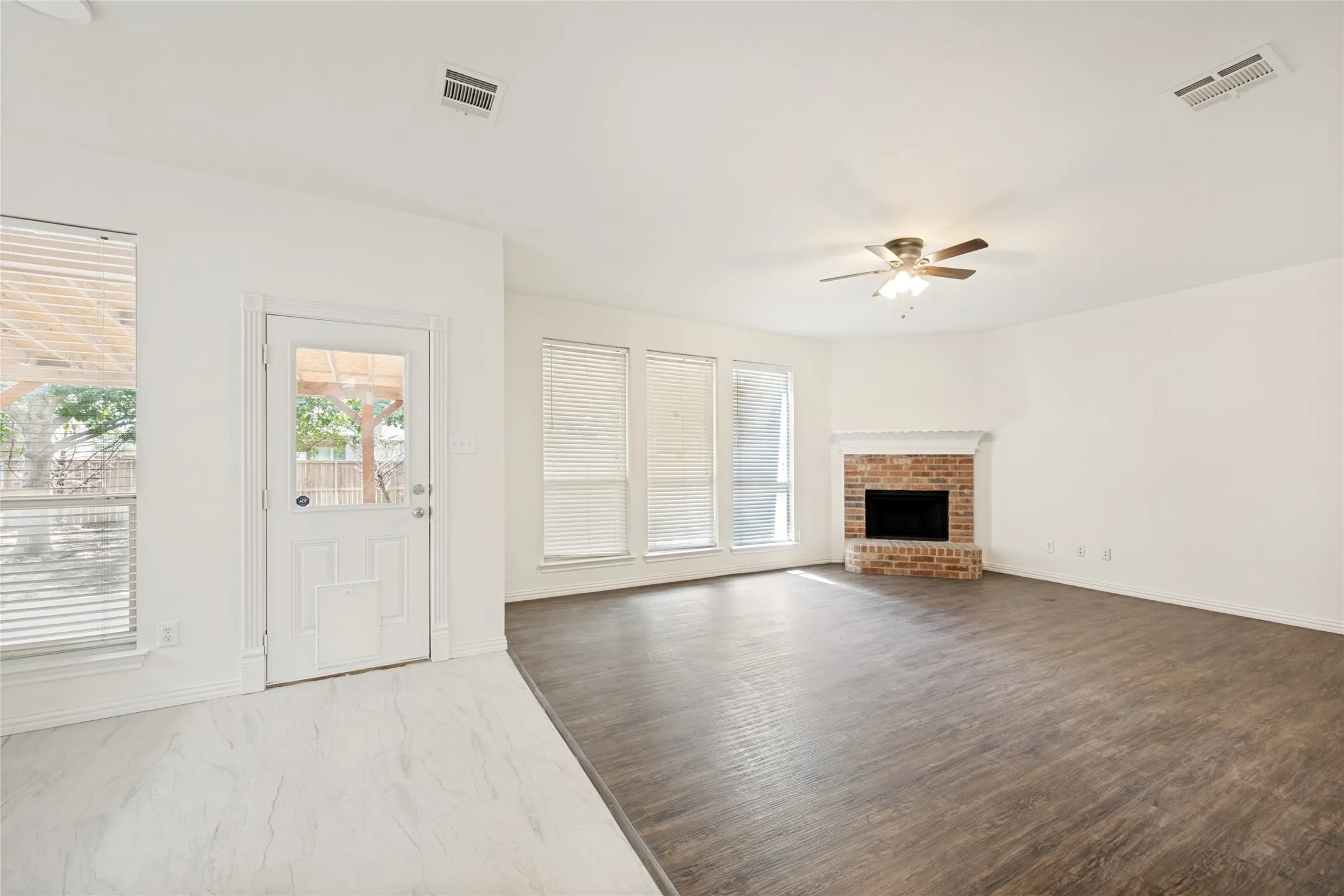 Spacious living area featuring a corner brick fireplace with a white mantel, ceiling fan, and large windows with blinds