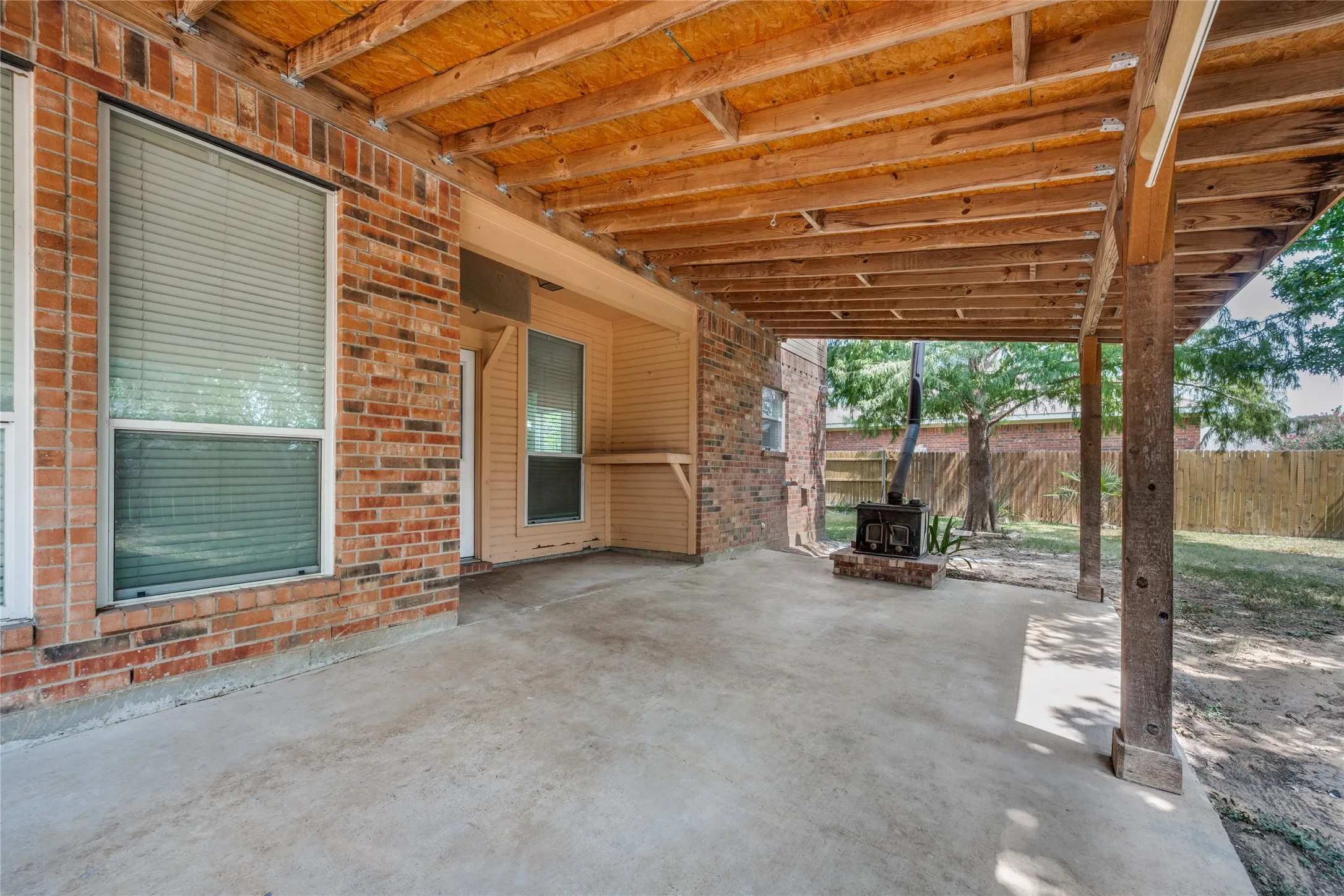Spacious covered patio featuring a concrete slab, exposed wood beam ceiling, brick exterior, and a built-in outdoor fireplace