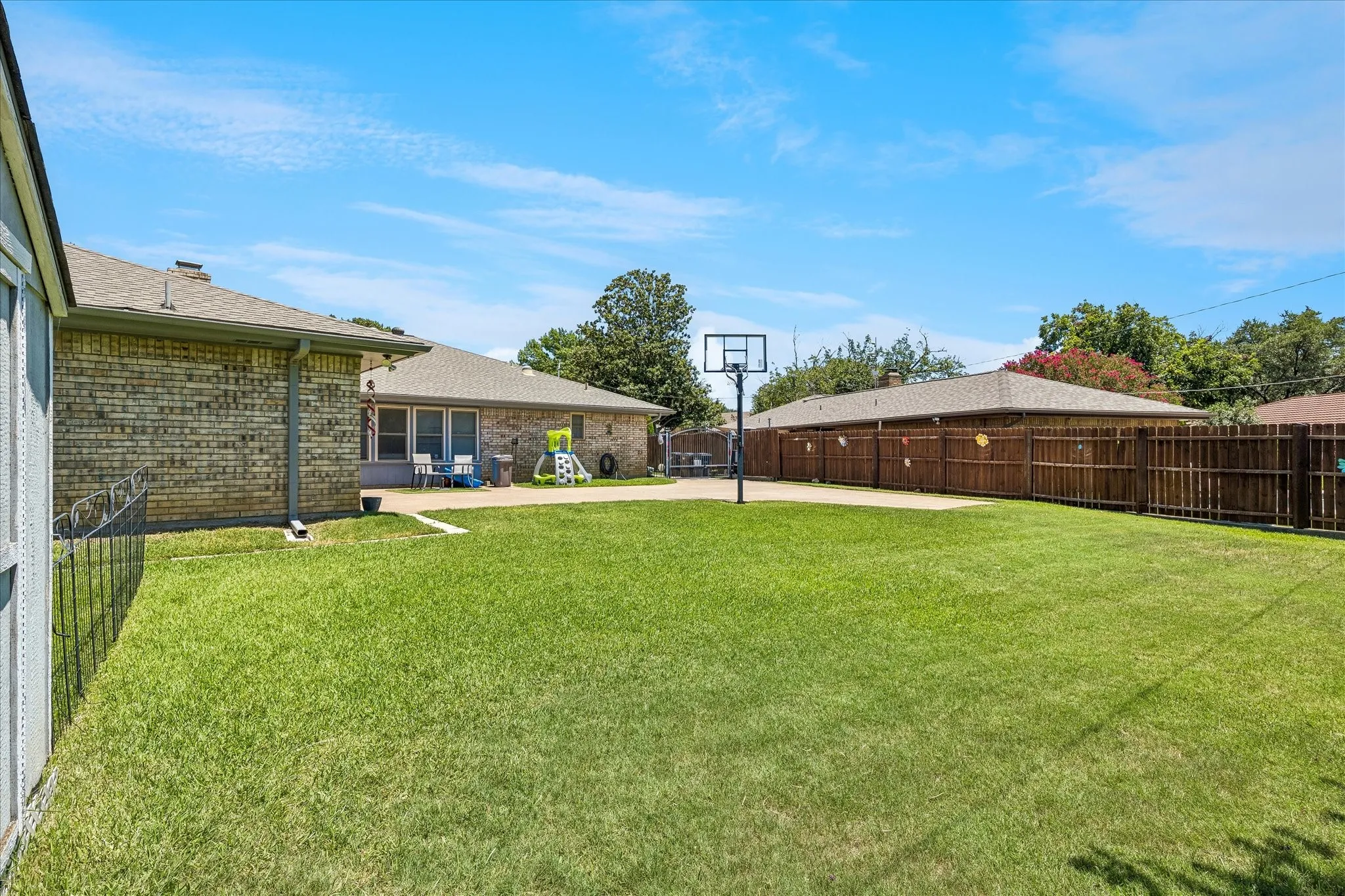 Expansive backyard featuring a manicured lawn, basketball hoop, mature landscaping, and wooden privacy fencing