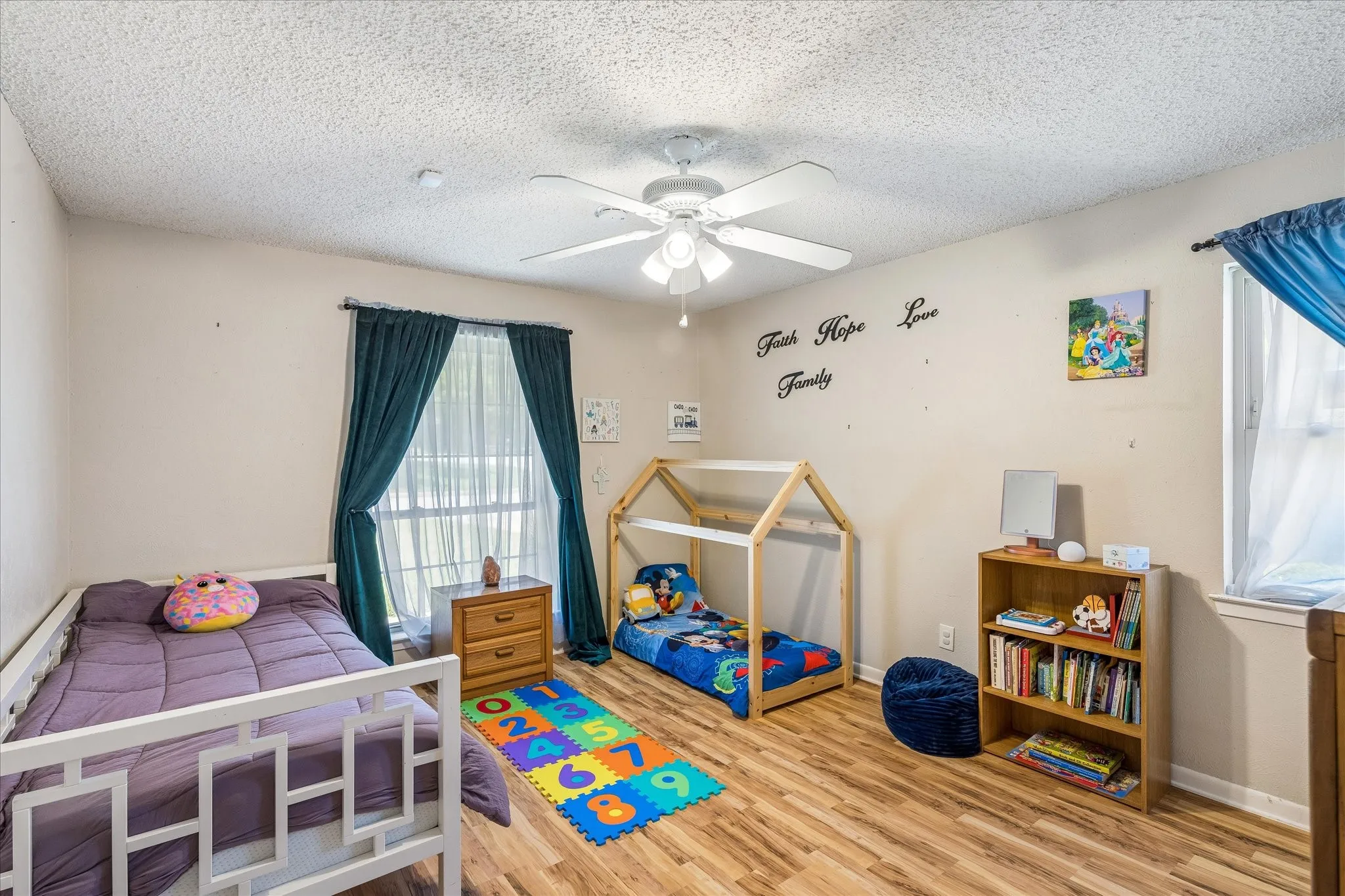 Room featuring wood-finish flooring, two windows with vertical blinds and dark curtains, a ceiling fan with integrated lighting, and light-toned walls