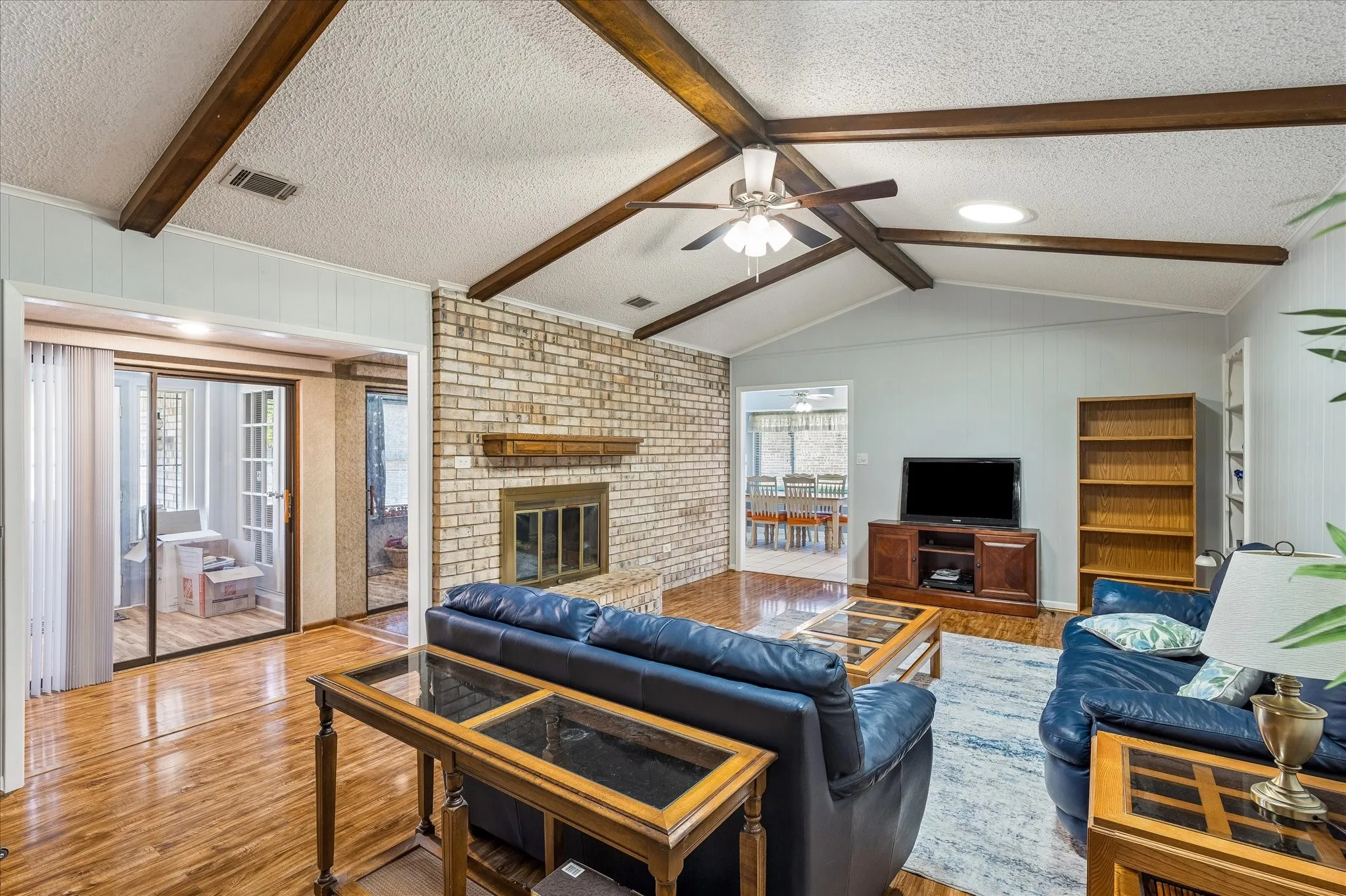 Living area with vaulted ceiling featuring exposed wood beams, brick fireplace with wood mantel, wood-finish flooring, and sliding glass doors