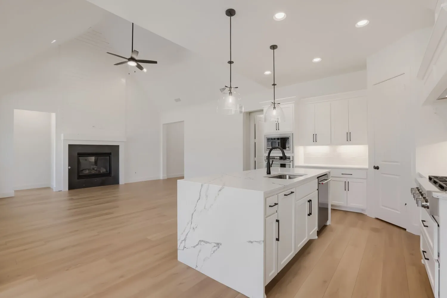 Vaulted ceiling with a black ceiling fan, black fireplace with a white mantel, wood-finish flooring, kitchen island with a waterfall countertop, and pendant lighting