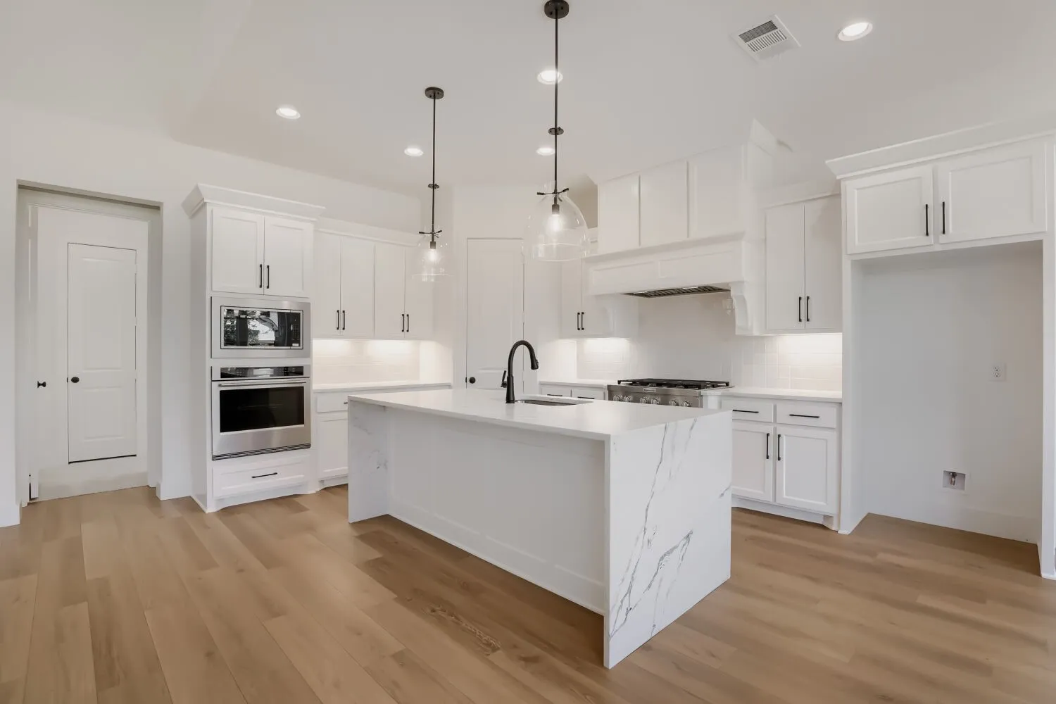 Contemporary kitchen featuring white shaker cabinetry, a large waterfall-edge island with quartz countertop, stainless steel appliances including dual ovens and gas cooktop, pendant lighting with metal fixtures, and warm wood-finish flooring throughout