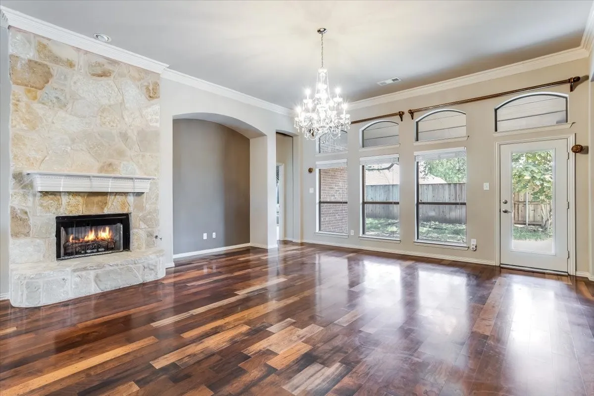 Elegant great room showcasing rich hardwood flooring in a herringbone pattern, a gas fireplace with natural stone surround, soaring ceilings with crystal chandelier, and expansive multi-pane glass doors and windows framing natural light throughout the space