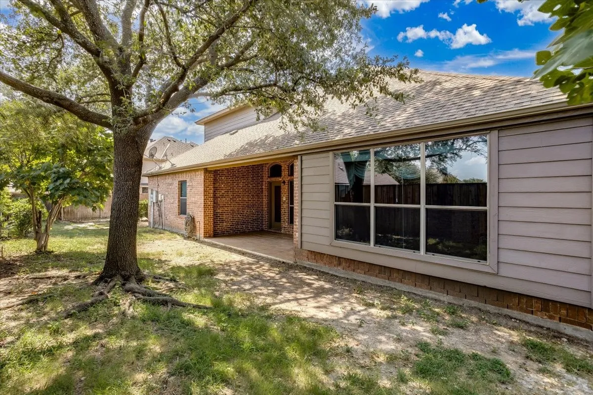 Spacious backyard featuring a prominent mature tree, a covered patio area with brick siding, and a large multi-pane window