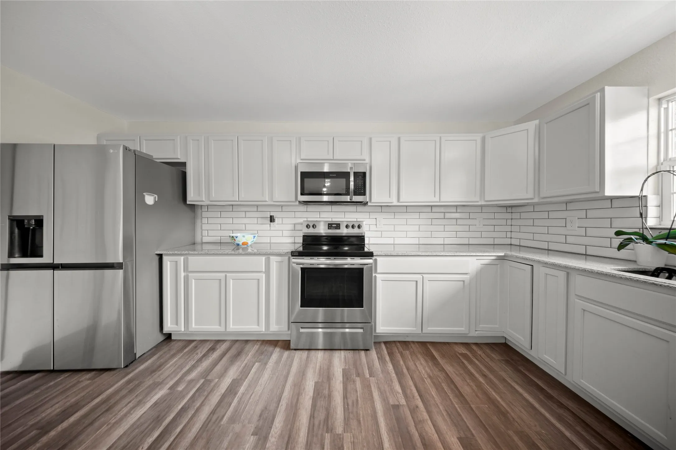 Kitchen featuring white shaker-style cabinetry, stainless steel appliances, speckled countertops, a white subway tile backsplash, and wood-finish flooring