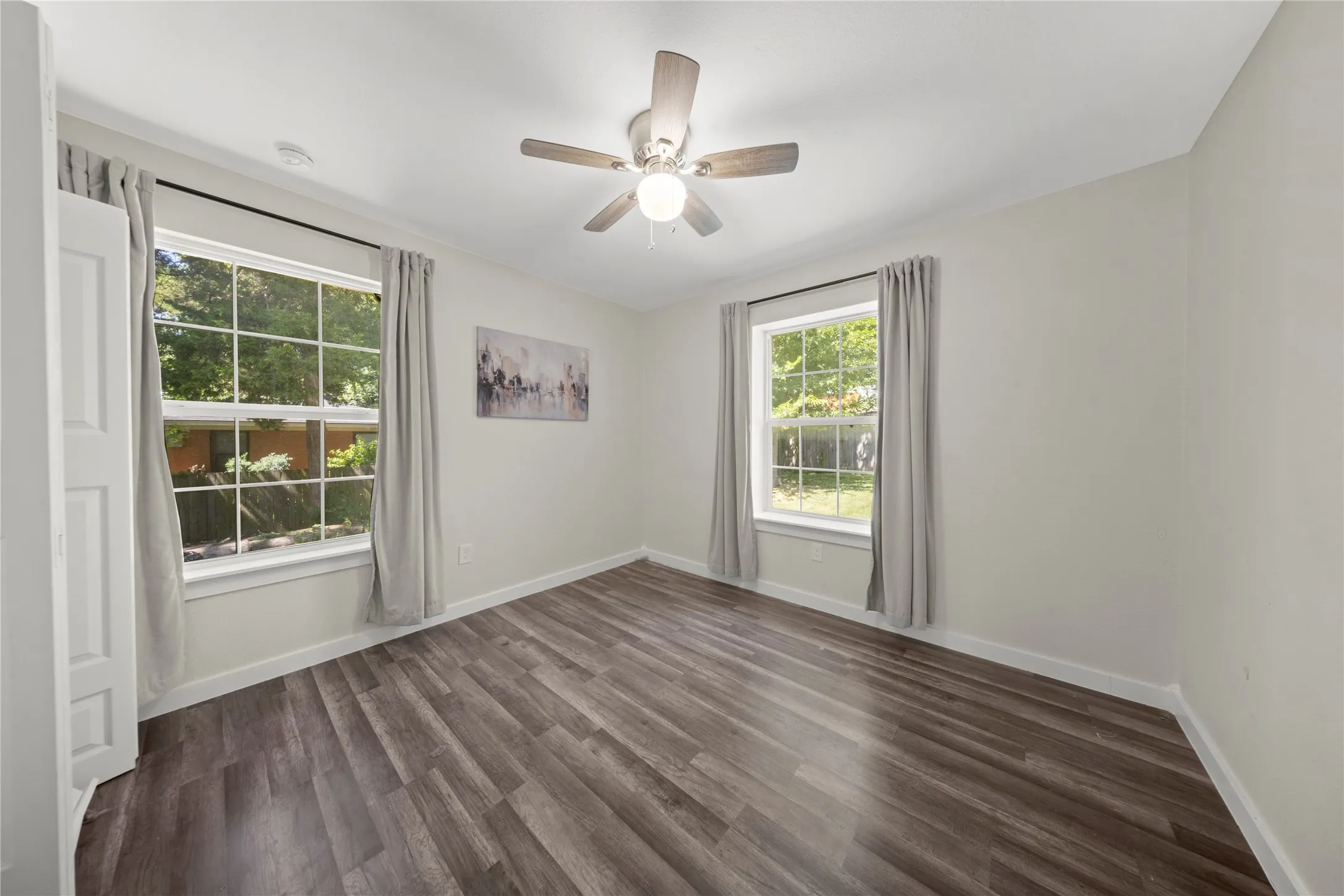 Spacious room featuring wood-finish flooring, two large windows with white trim, and a ceiling fan with integrated lighting
