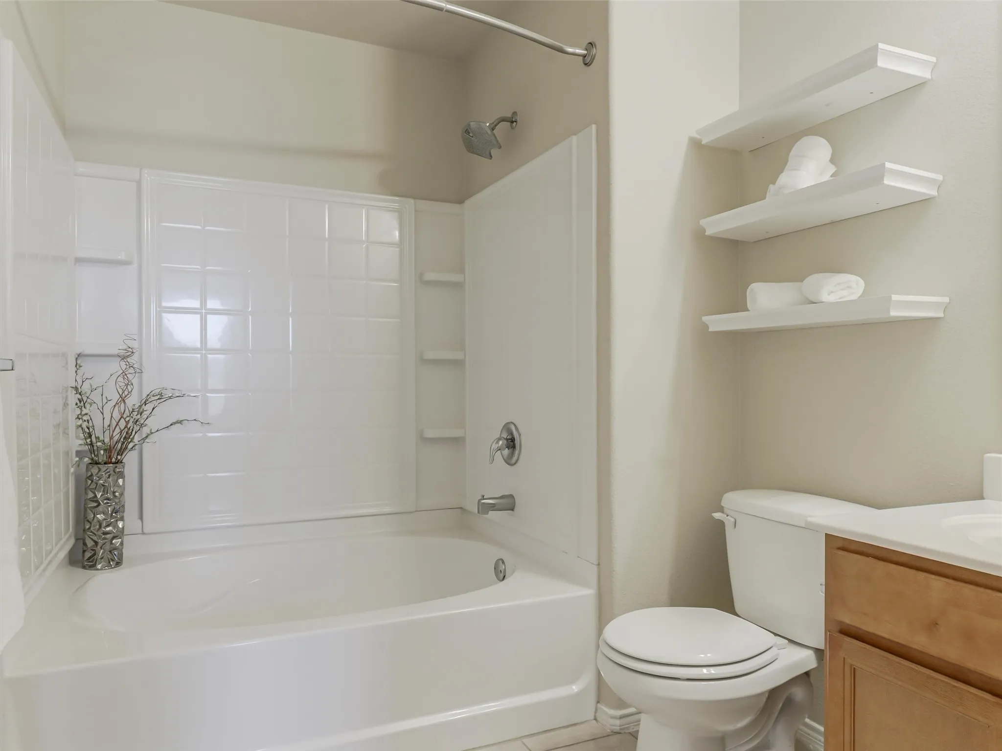 White tiled bathtub with a shower fixture, wall-mounted shelving, and a toilet