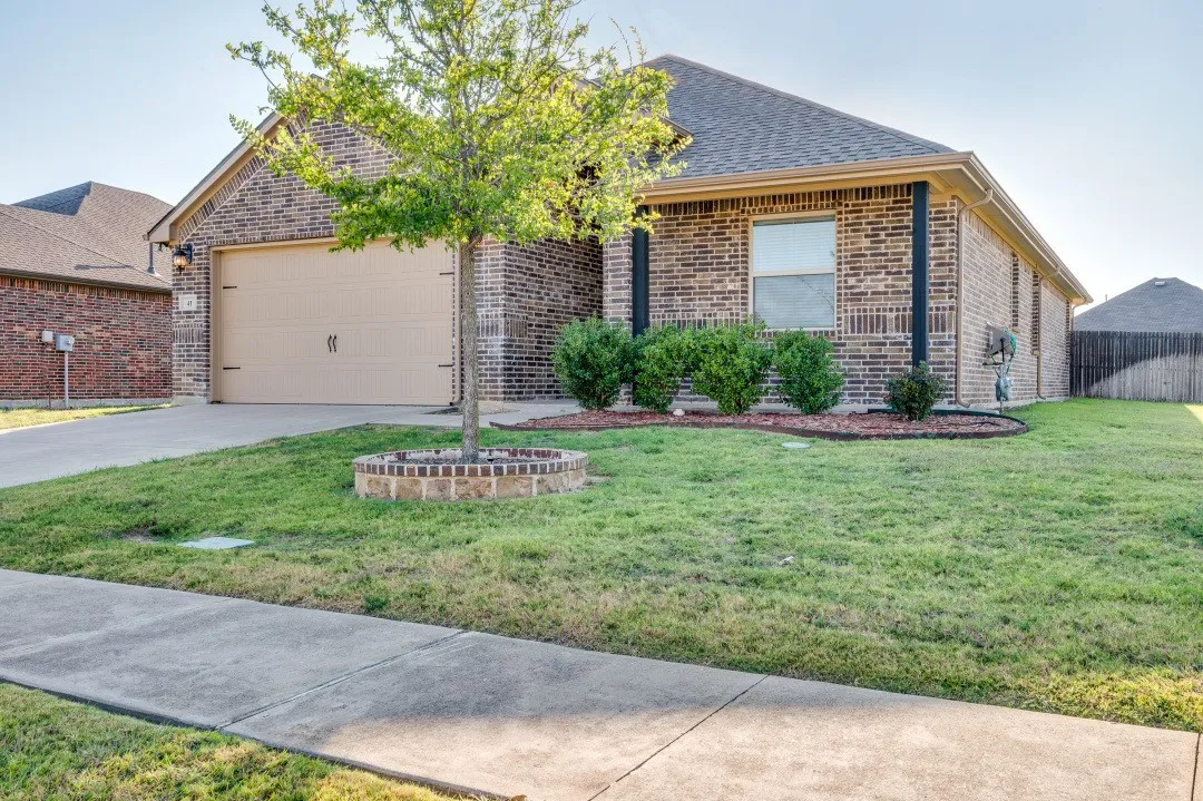 Brick exterior with an attached garage, paved driveway, and established landscaping with shrubbery