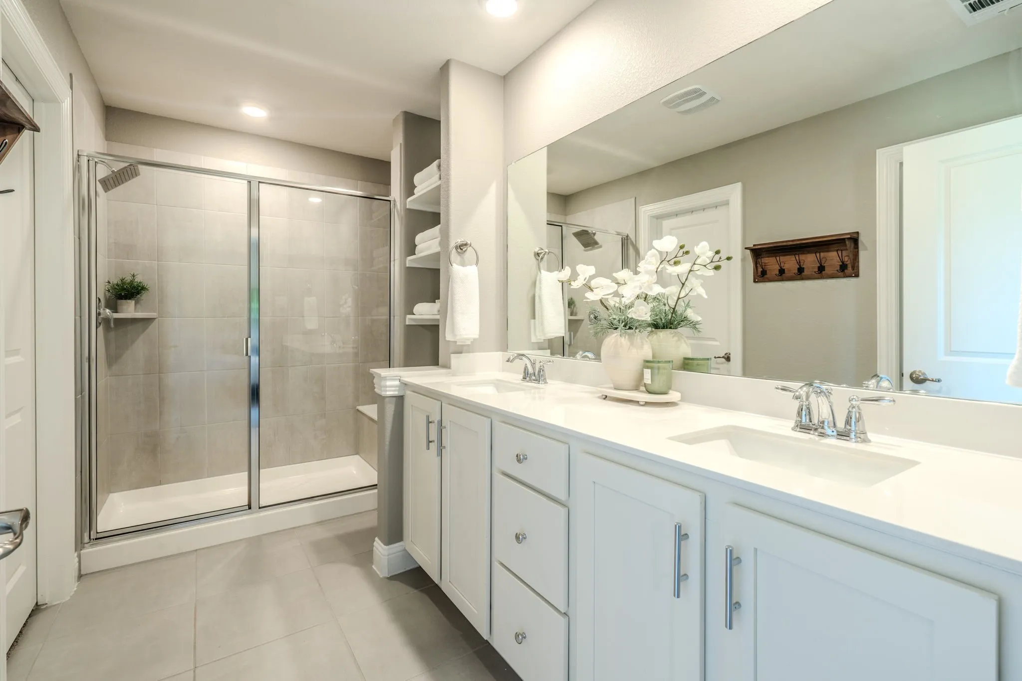 7329 Allium Court, Little Elm, TX 76227 - Primary Bathroom - Dual sink vanity with white cabinetry and chrome hardware, a full-width mirror, a glass-enclosed shower with a built-in bench, and recessed shelving