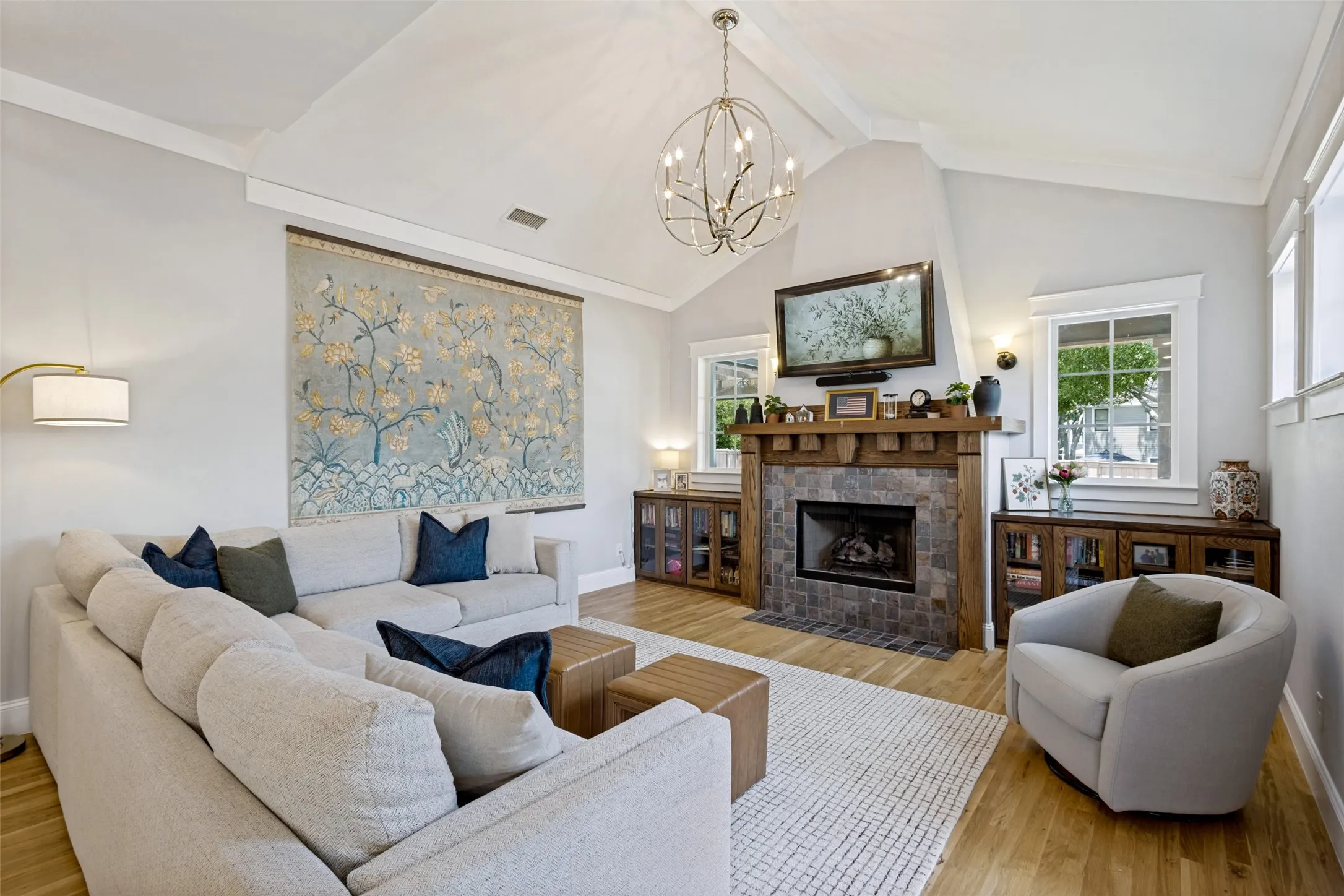 Vaulted ceiling with a contemporary chandelier, stone-faced fireplace with a wood mantle, wood-finish flooring, built-in cabinetry, and multiple windows