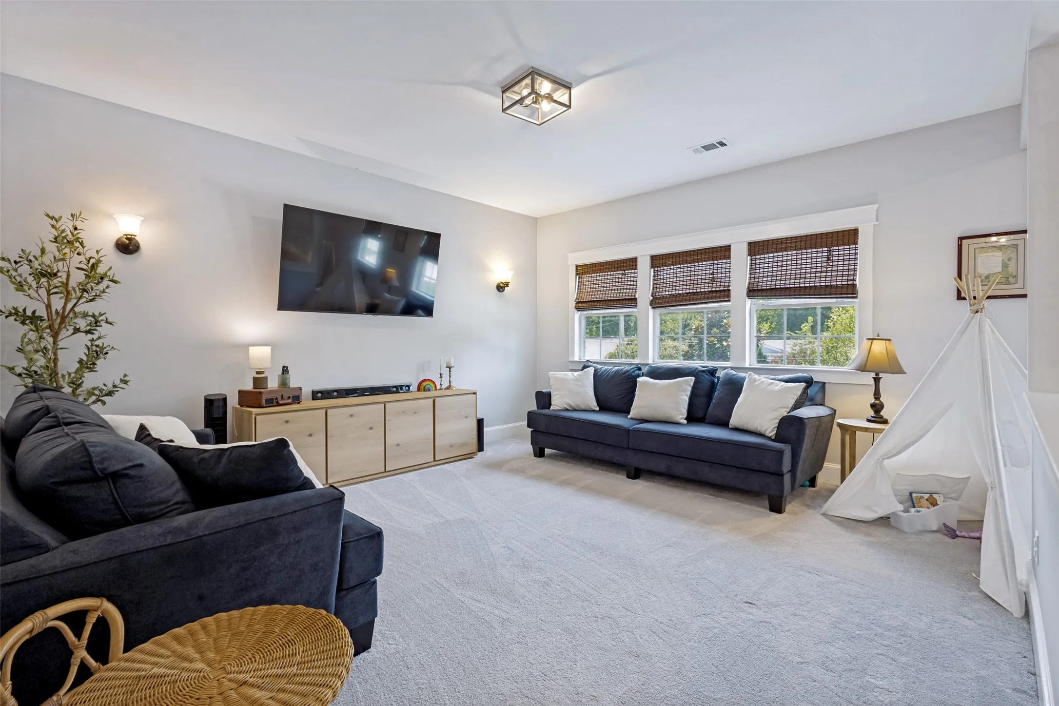 Wall-mounted television with a light wood-finish media console, three windows with natural woven shades, ceiling-mounted fixture, and sconce lighting