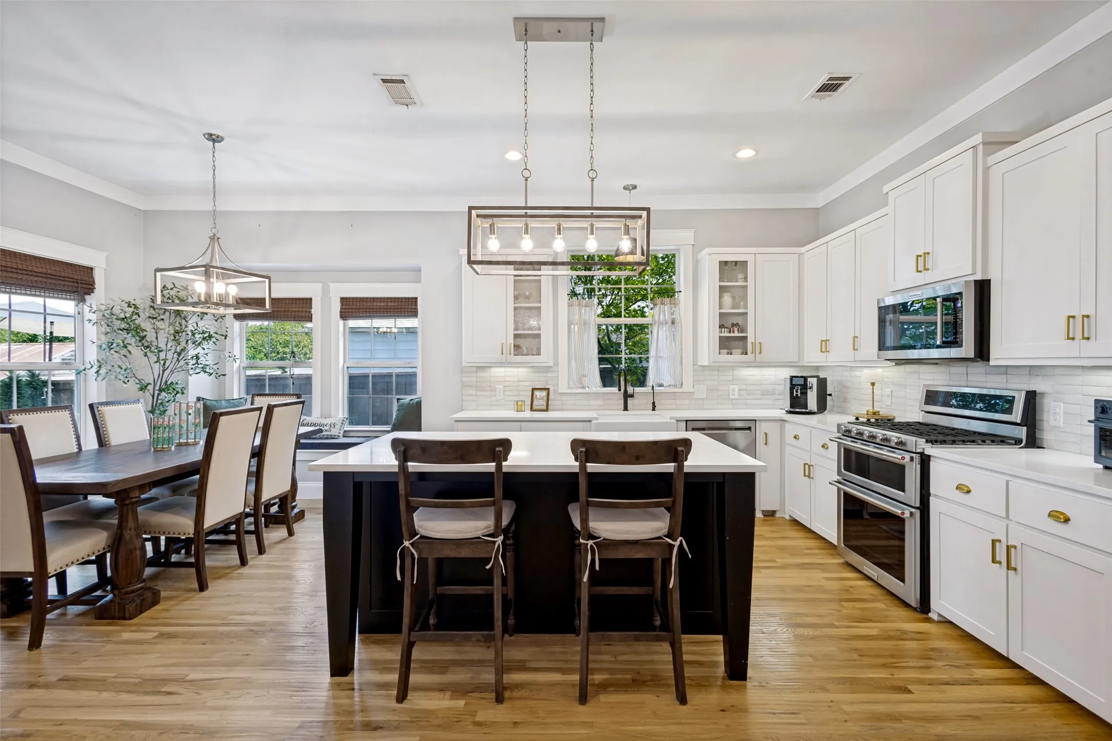 Open-concept living area featuring wood-finish flooring, a kitchen island with seating, pendant lighting, white cabinetry with gold-tone hardware, and stainless steel appliances