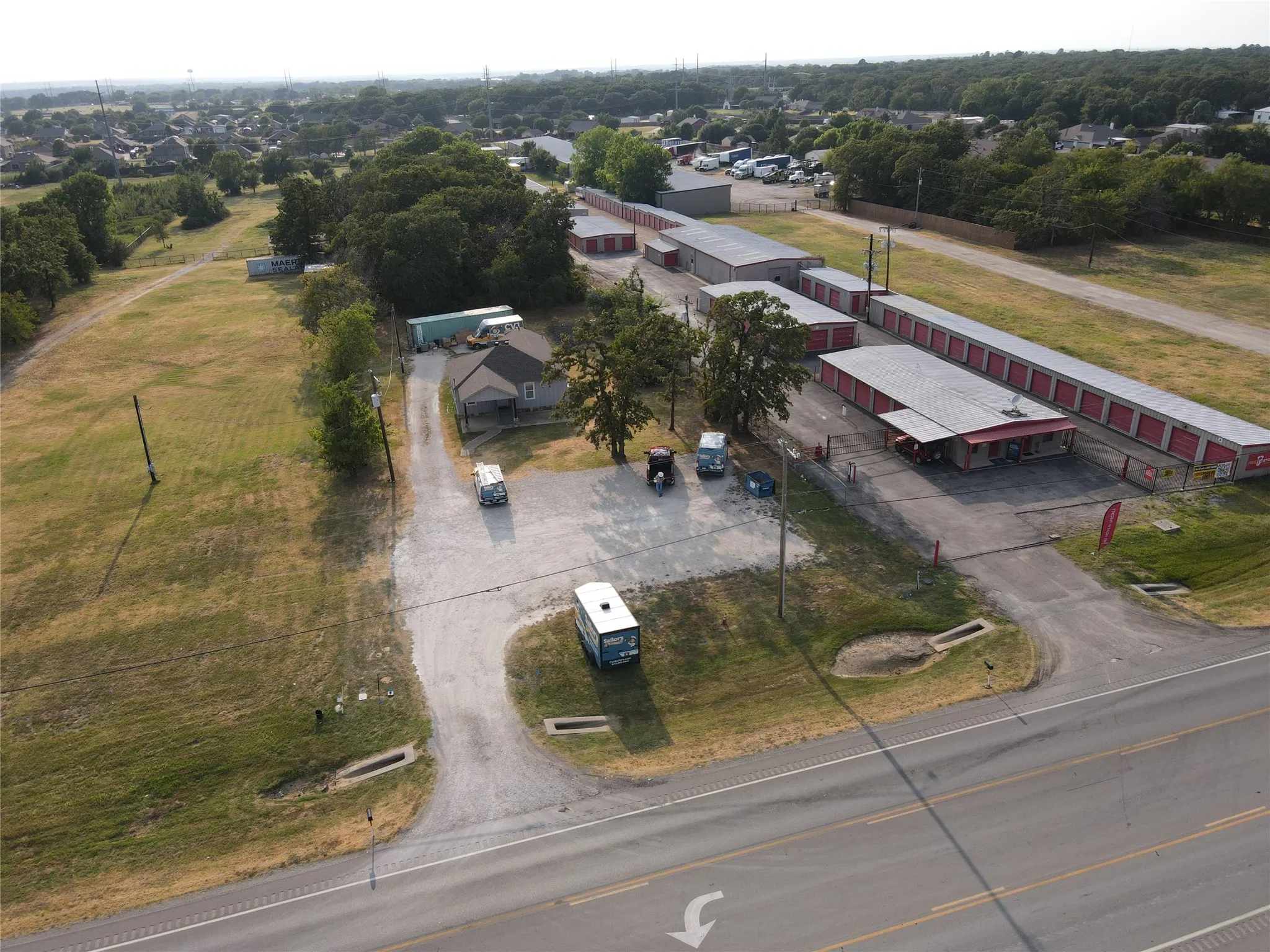 Aerial perspective showcasing a commercial property with multiple storage unit buildings, a central office structure, and extensive gravel parking areas