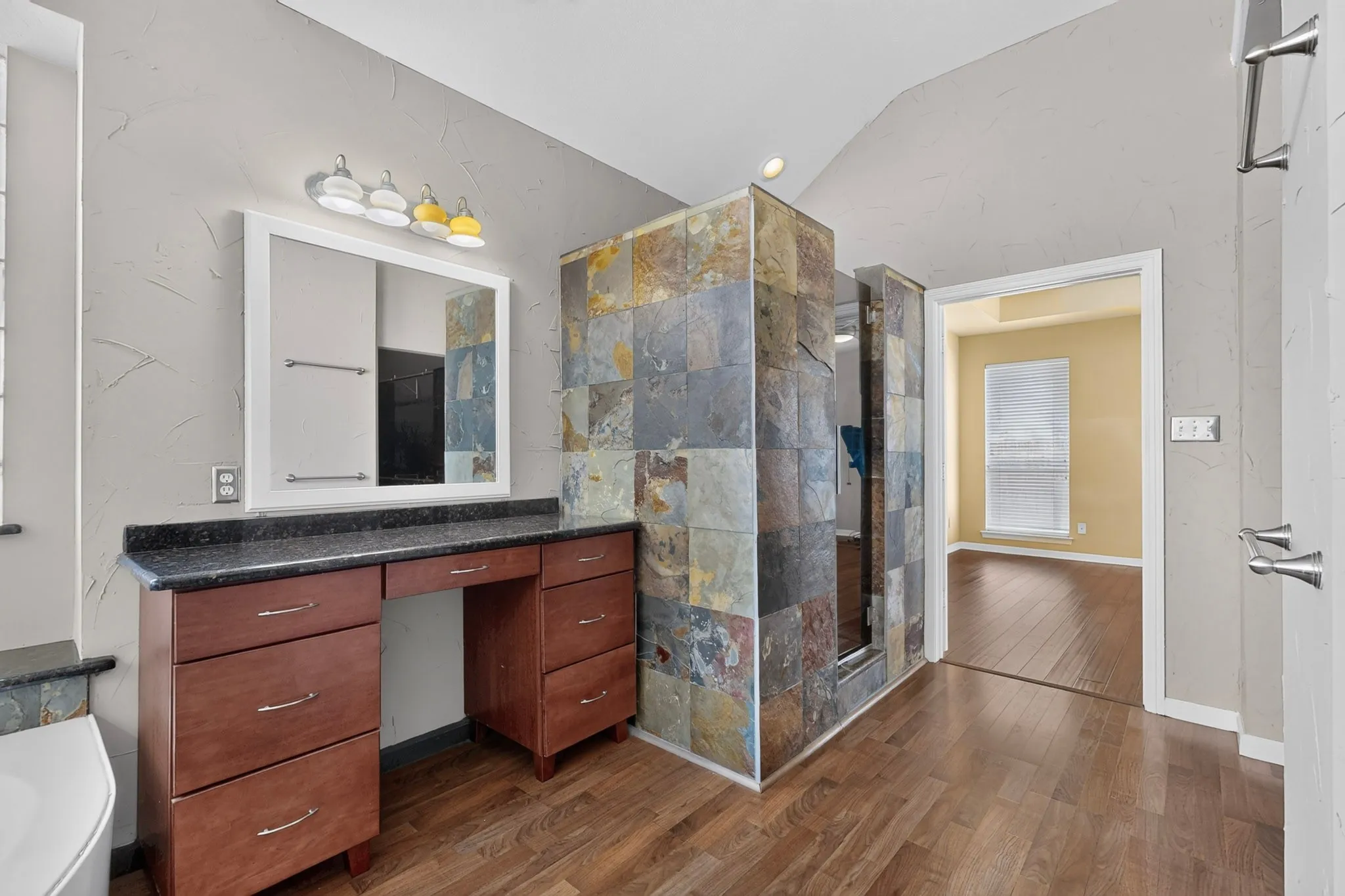 Wood-finish flooring, vanity with built-in cabinetry, dark countertop, framed mirror, and a tiled shower enclosure