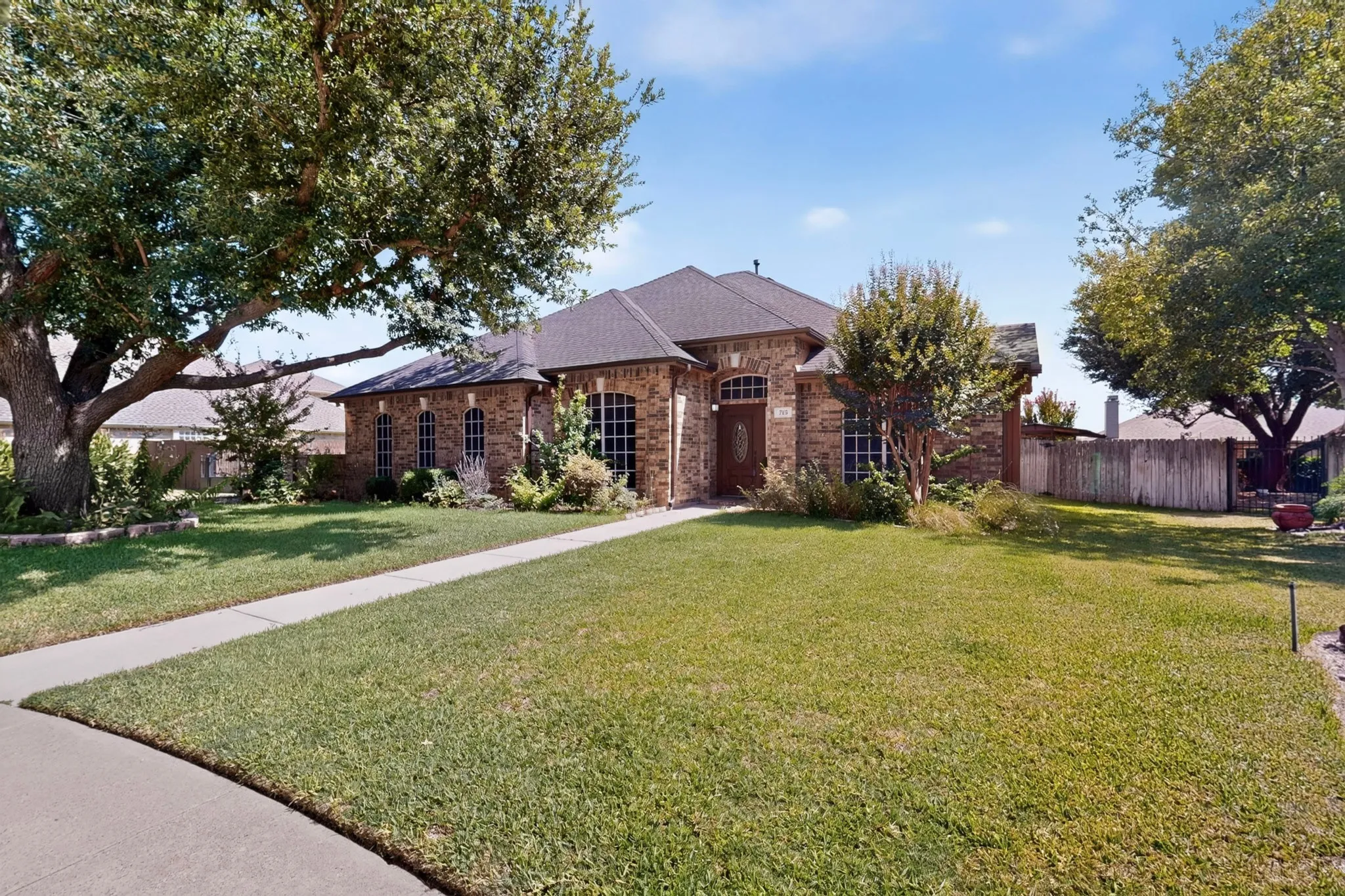 Brick exterior featuring arched windows, a dark shingle roof, mature trees, and a concrete walkway