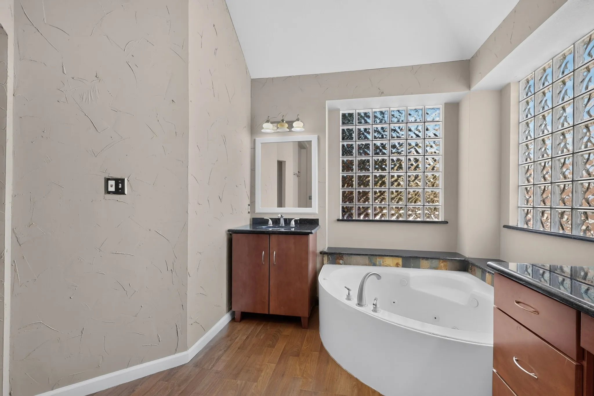 Soaking tub with deck-mounted fixtures, two glass block windows, slate-style tile surround, wood-finish flooring, and a vanity with dark countertop