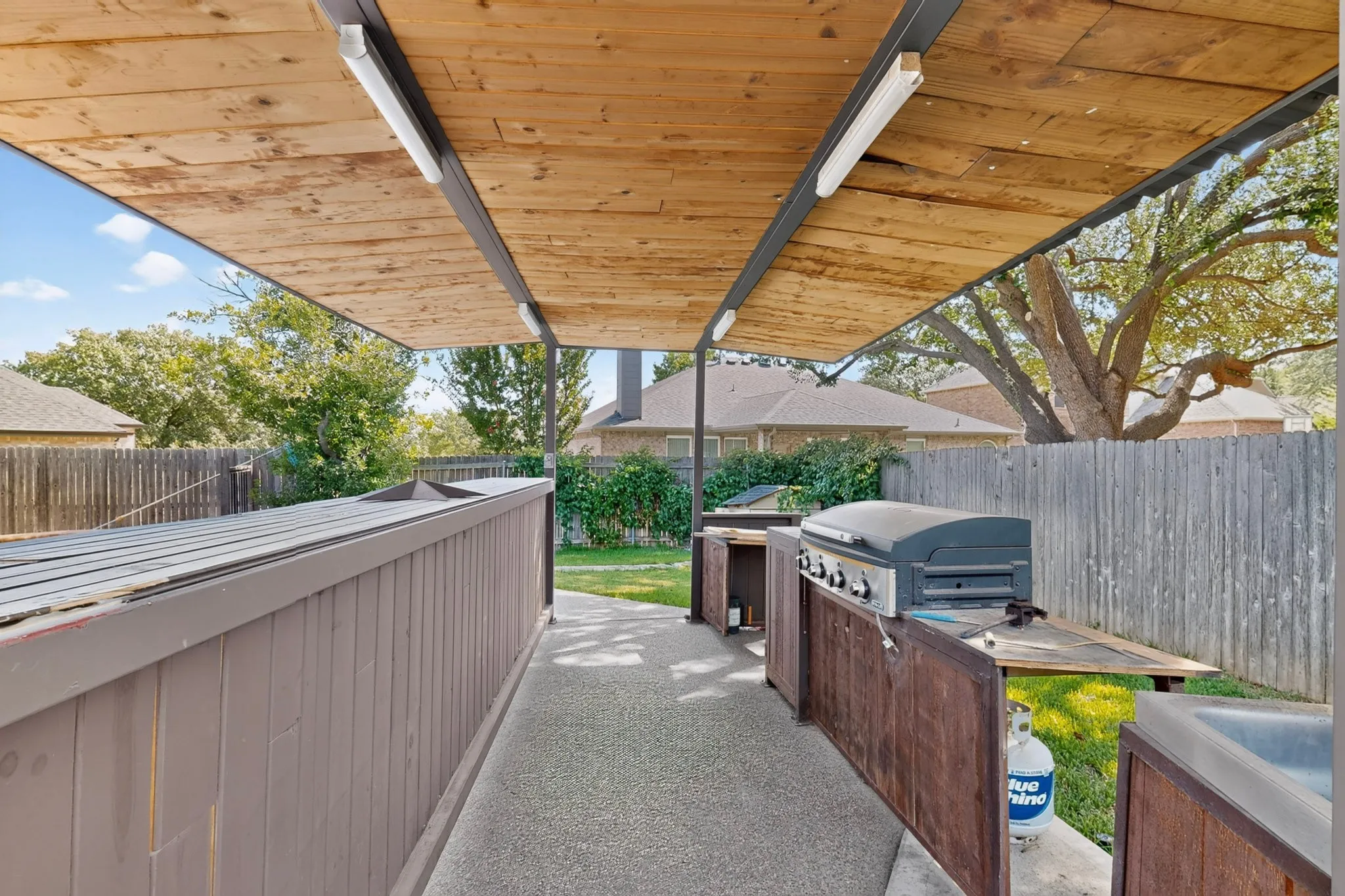 Covered outdoor kitchen featuring a built-in grill, integrated sink, and an expansive wood-slat ceiling with recessed lighting