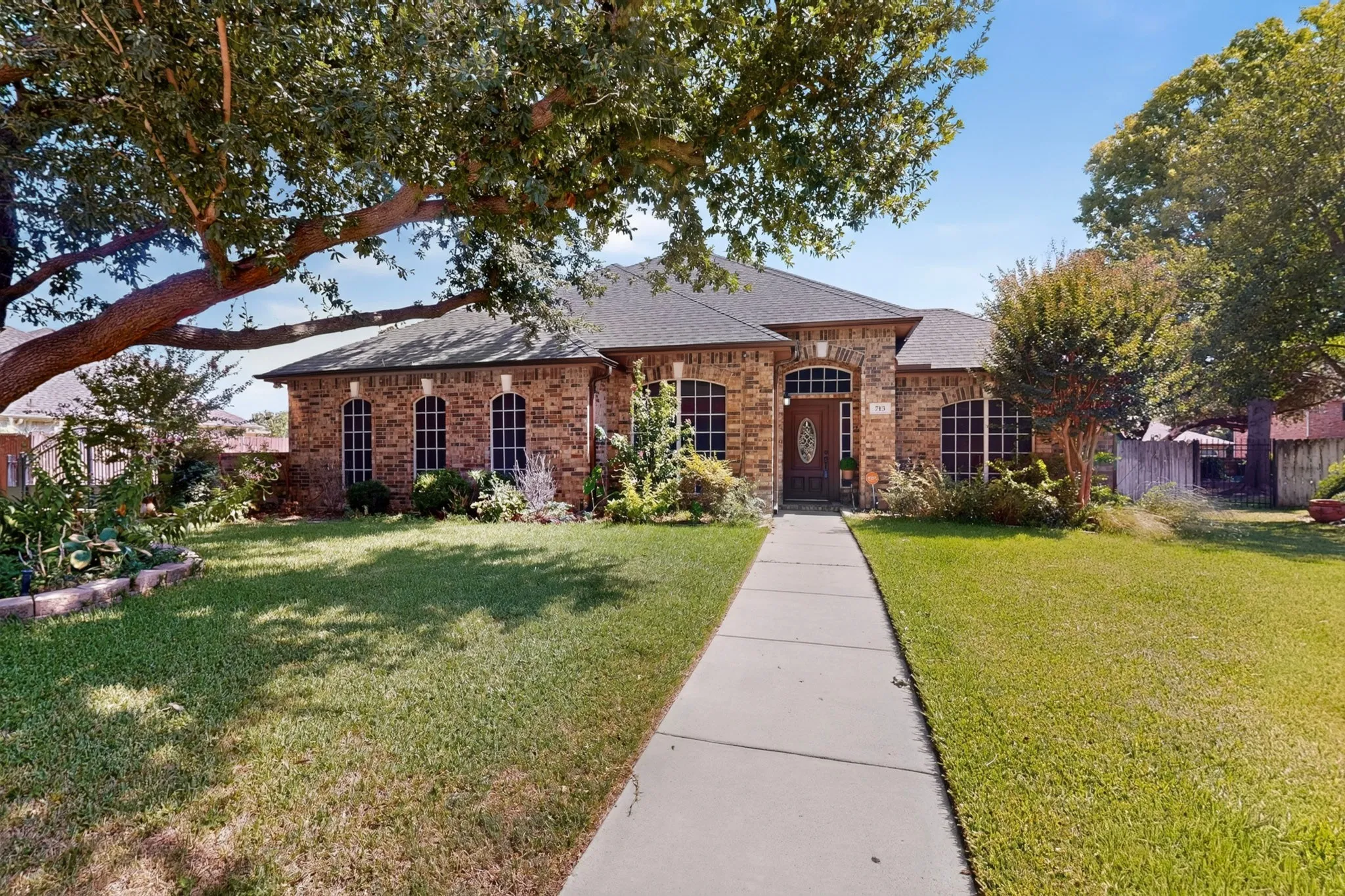 Brick exterior with a dark-pitched roof, arched windows, an ornate front door, a concrete walkway, and mature trees