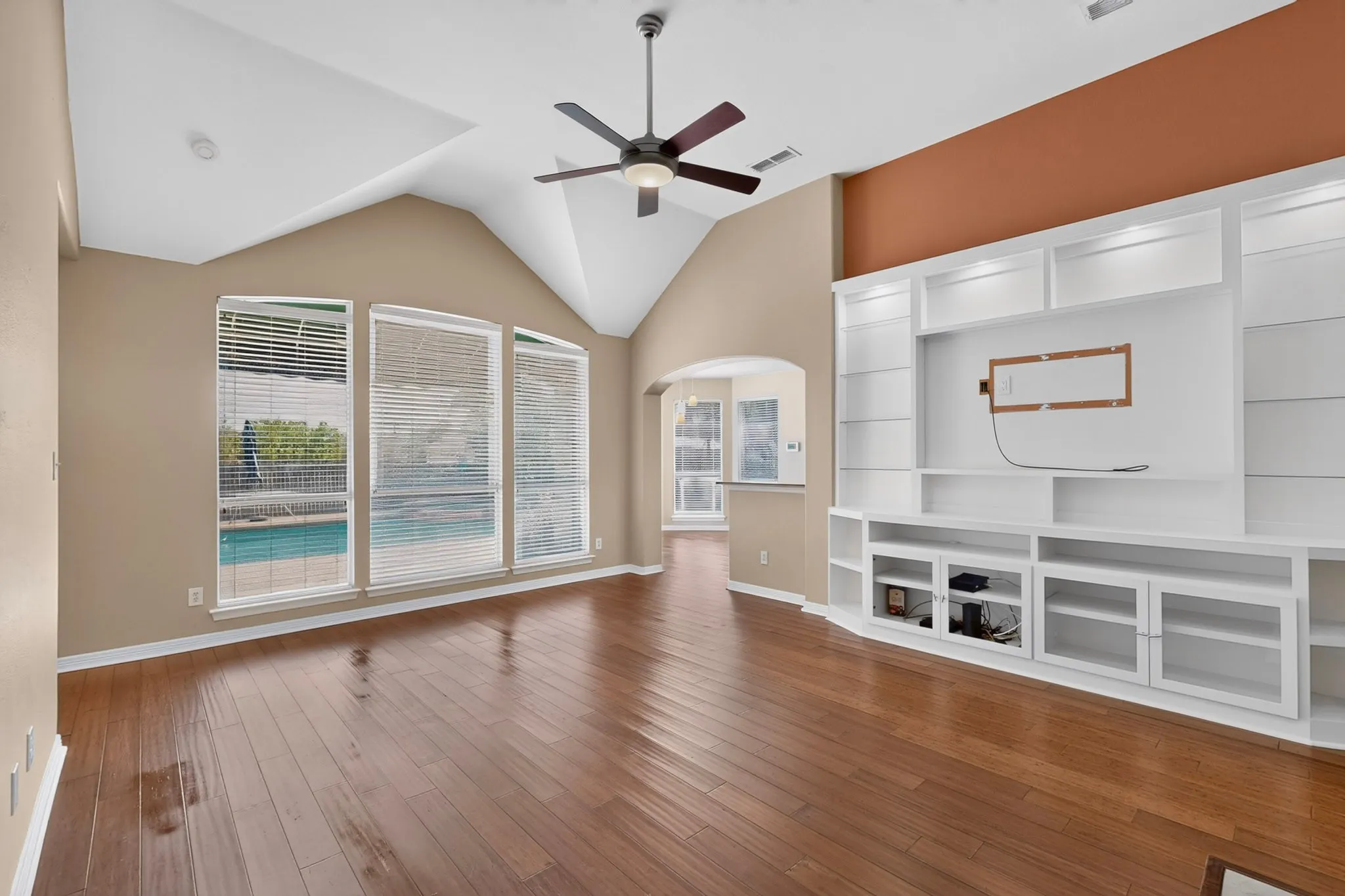 Vaulted ceilings, wood-finish flooring, custom built-in entertainment center, ceiling fan, and multiple windows overlooking a pool