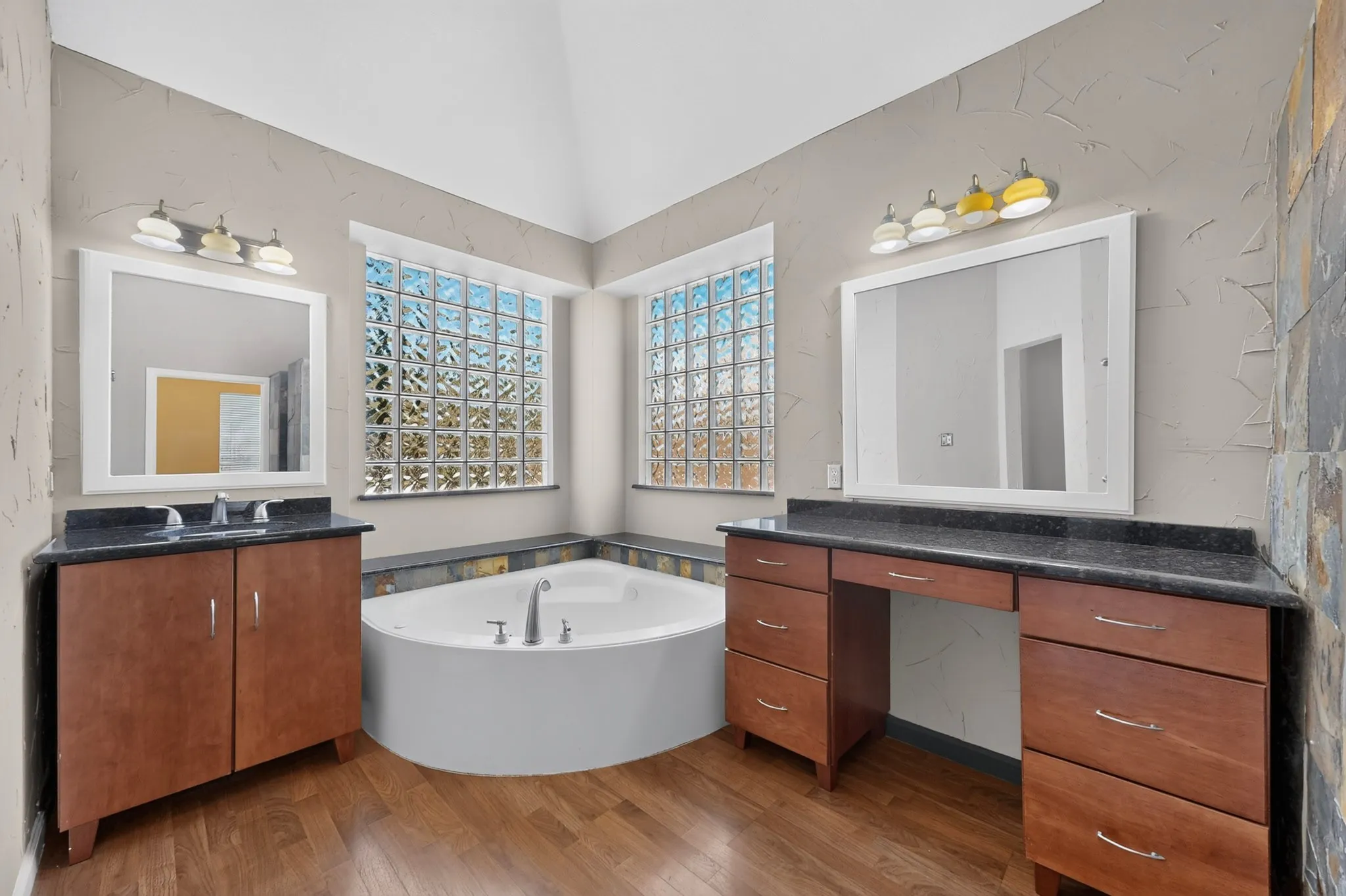 Soaking tub with deck-mounted fixtures, dual vanity sinks with dark countertops, wood-finish flooring, and glass block windows
