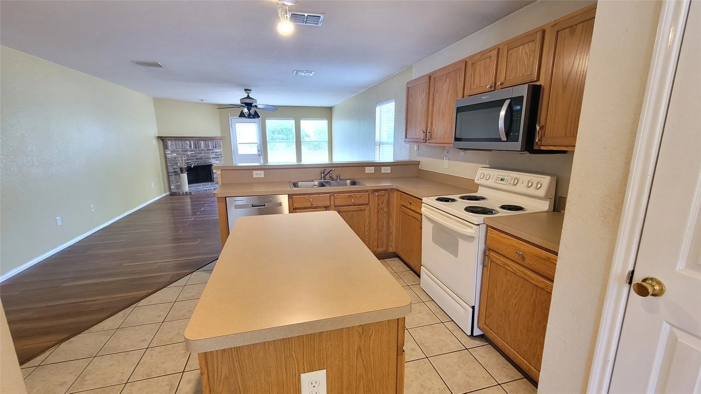 Kitchen with wood-finish cabinetry, a central island, and tile flooring, opening to a living area with wood-finish flooring and a stone-clad fireplace