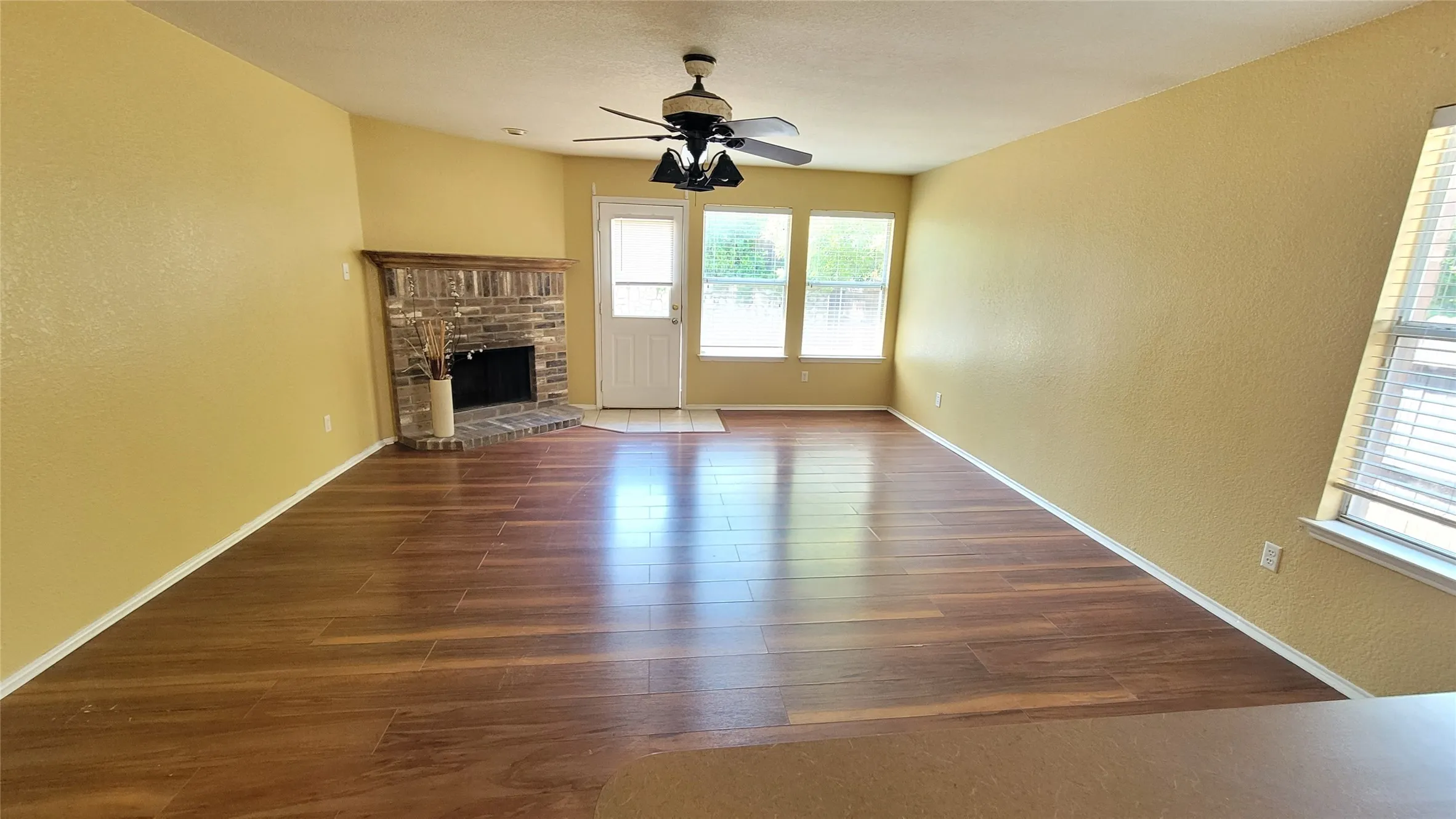 Spacious living area featuring wood-finish flooring, a brick fireplace, a ceiling fan, and multiple windows