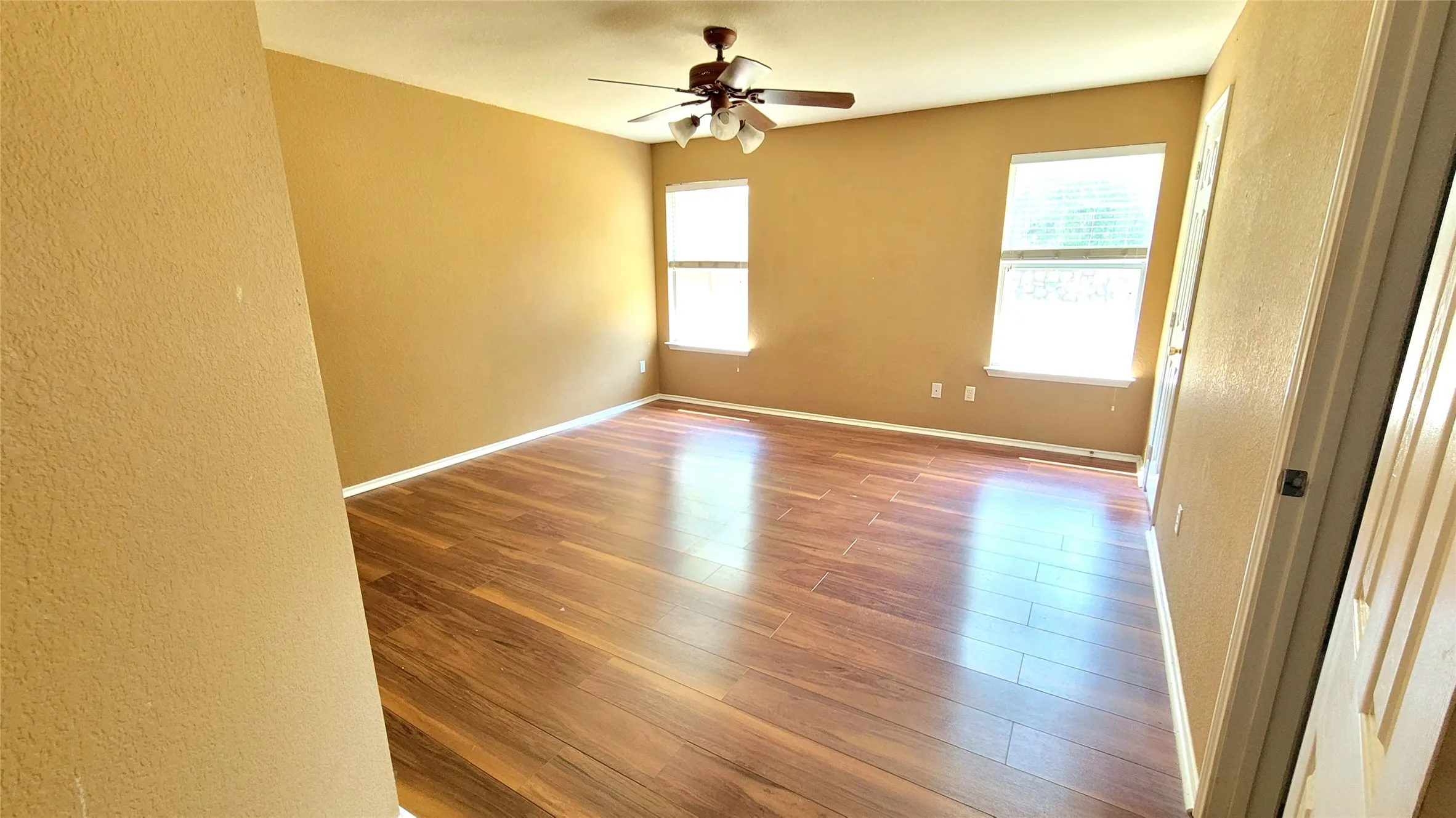 Spacious room featuring wood-finish flooring, two windows with blinds, a ceiling fan, and neutral-toned walls