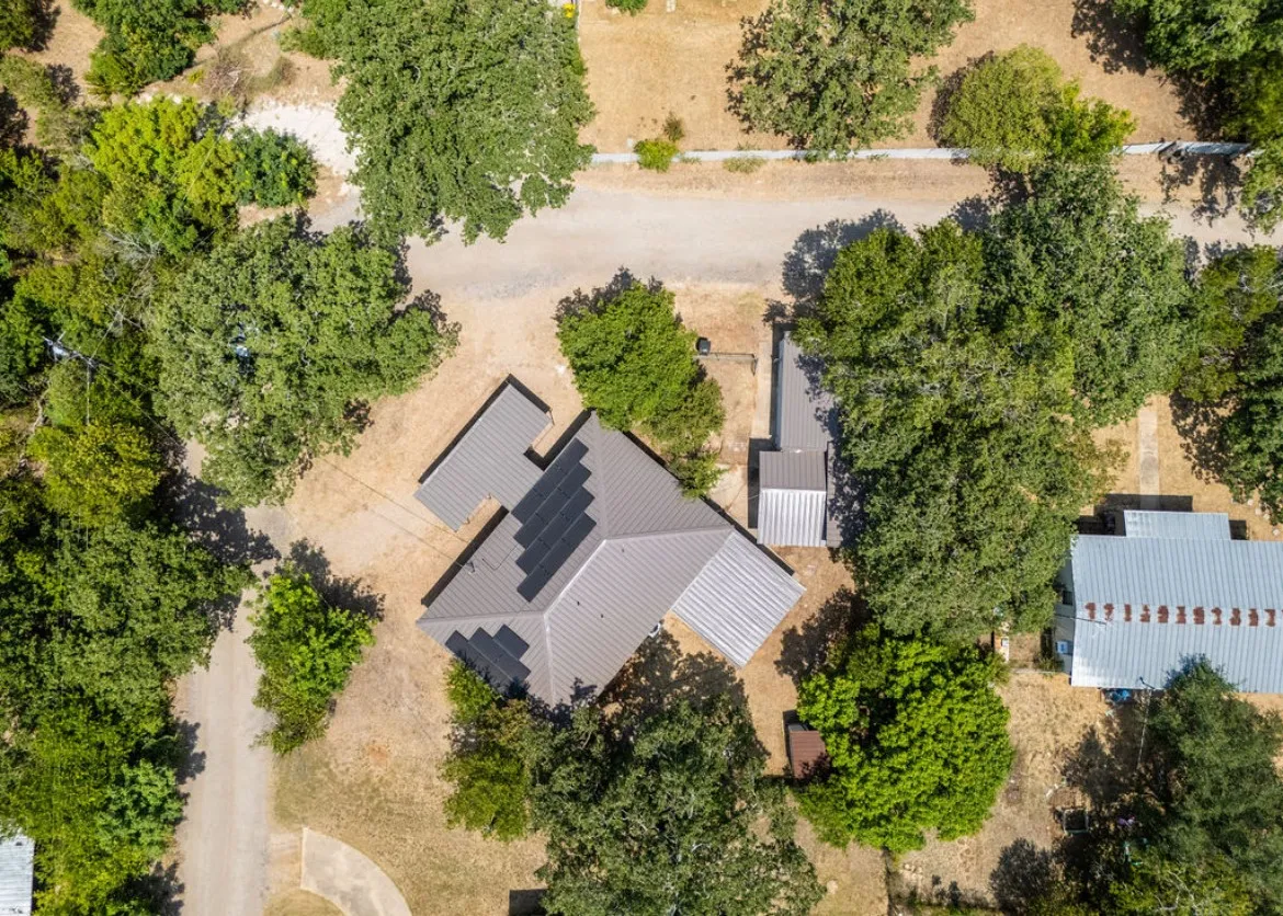 Aerial perspective showcasing a property surrounded by mature trees, featuring a main residence with a metal roof and an integrated solar panel array