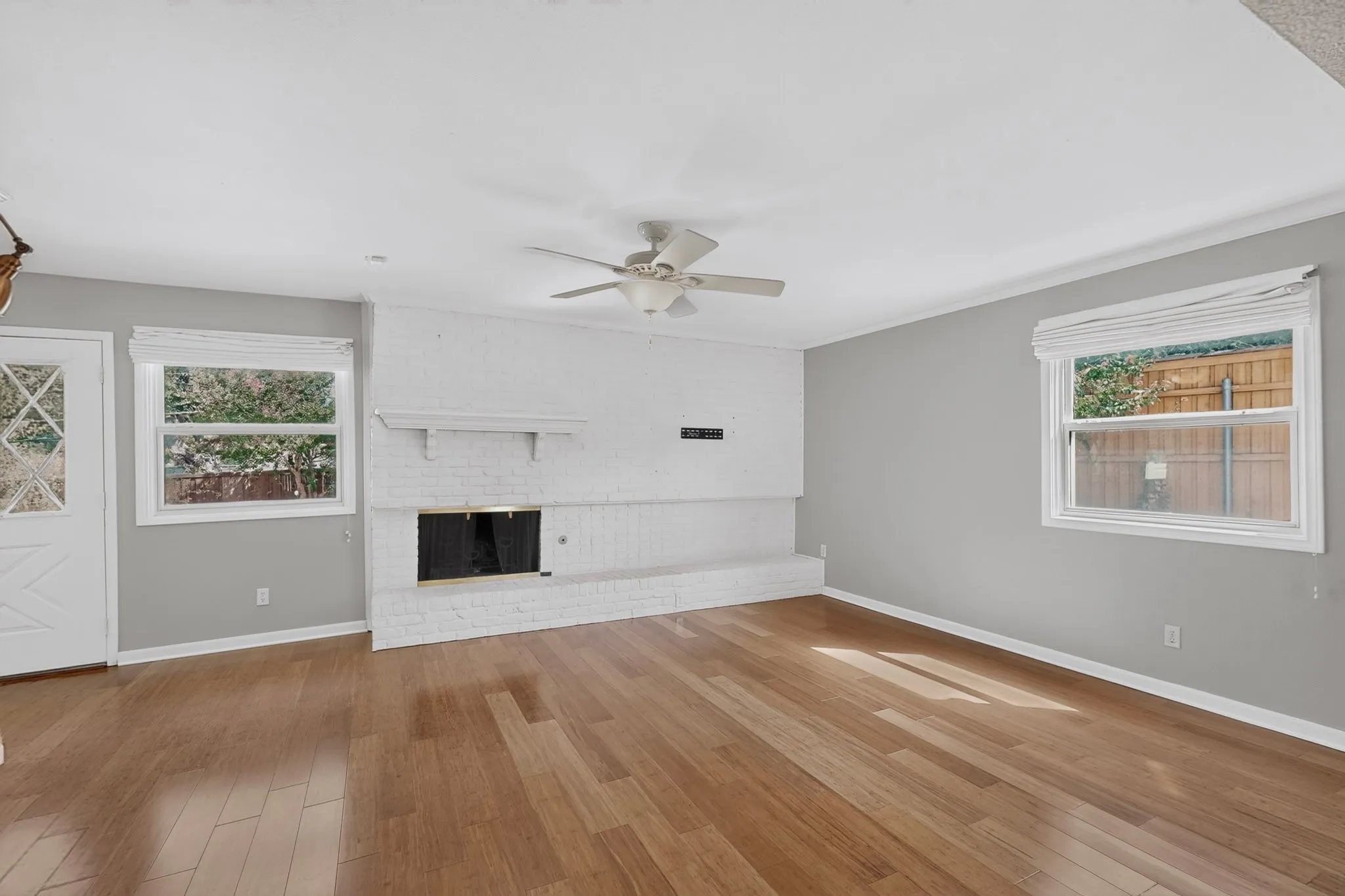 Wood-finish flooring, white brick fireplace with mantel, ceiling fan, white interior door with decorative glass, and multiple windows