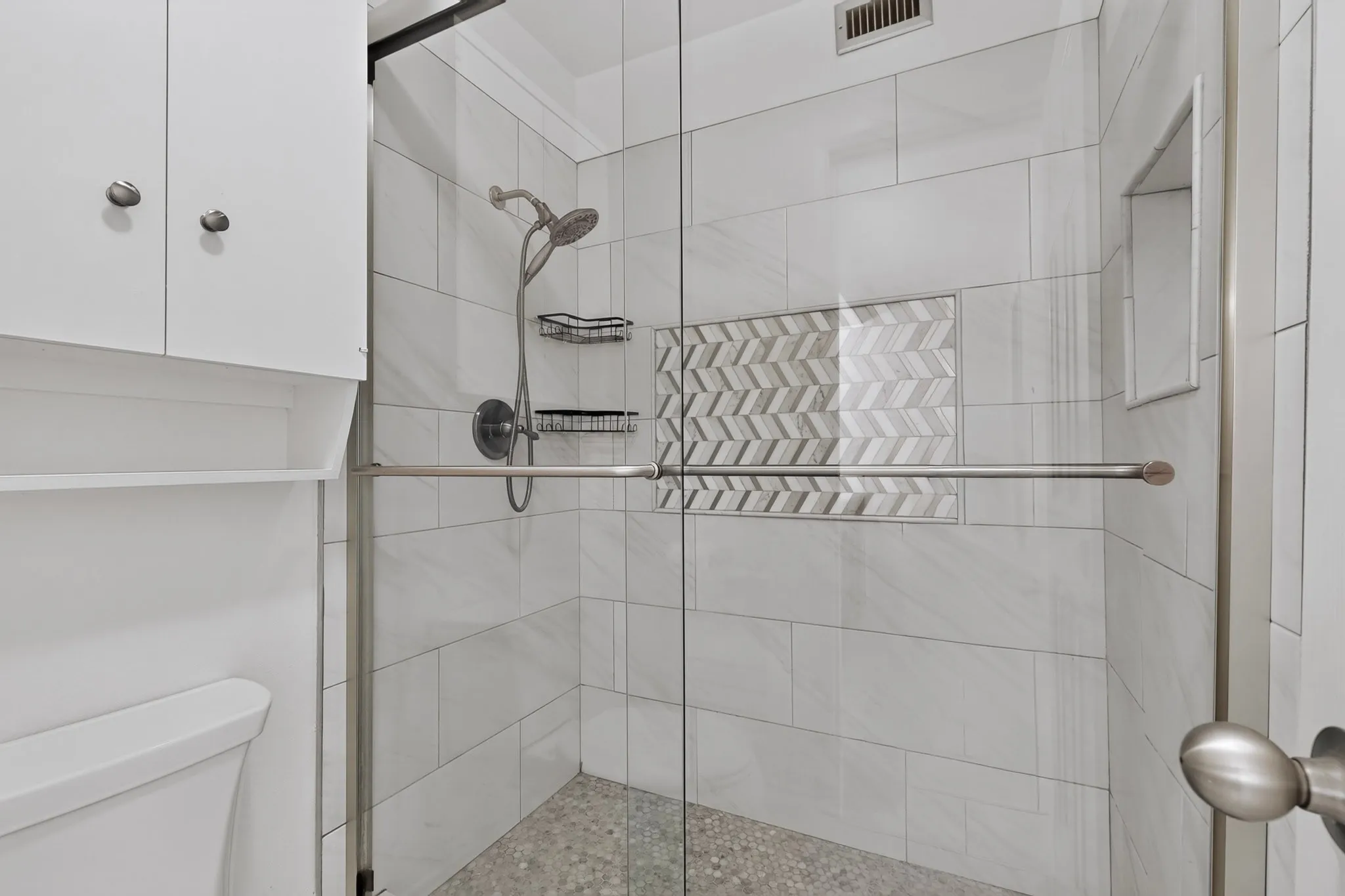 Walk-in shower with clear glass enclosure, subway-style tile surround, herringbone accent tile, integrated shower niche, and pebbled floor