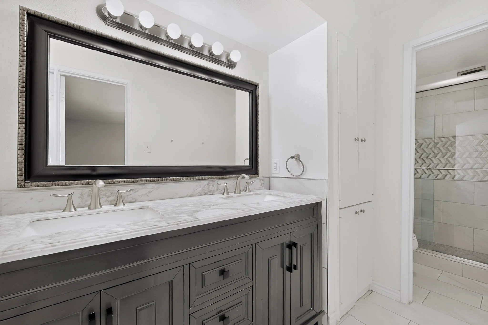 Dual undermount sinks set in a stone countertop, dark wood-finish vanity with ample storage, large framed mirror with overhead vanity lighting, white built-in storage cabinet, and a glass-enclosed shower with decorative tile inlay