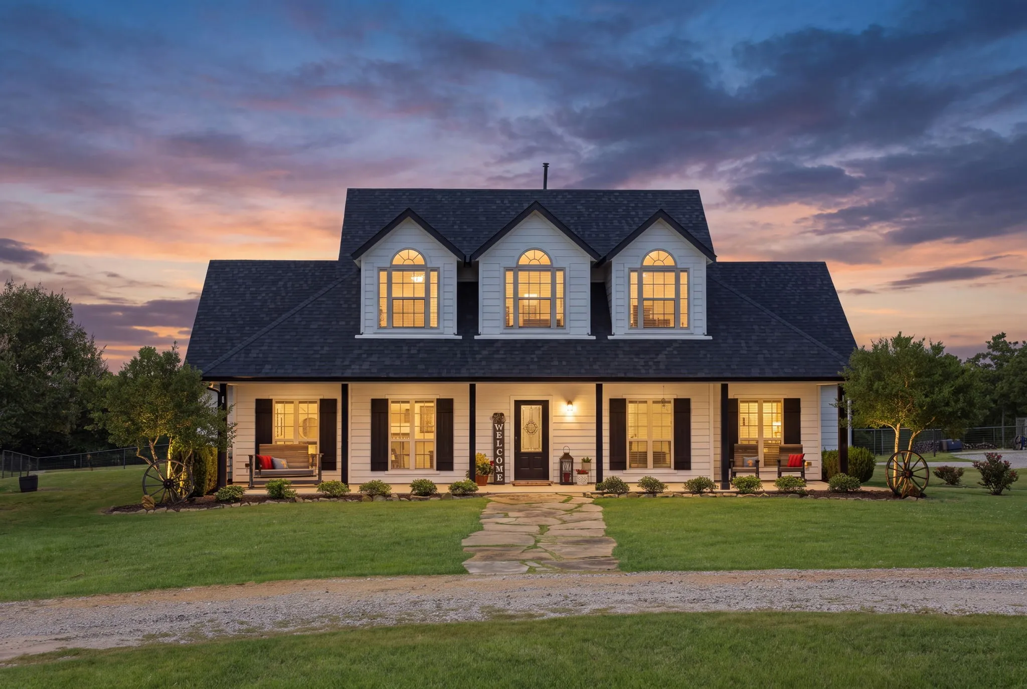 Glowing windows and three charming dormers welcome you home beneath a colorful twilight sky, while the expansive covered front porch and stone walkway set the scene for relaxed country living on 10.4 scenic acres.