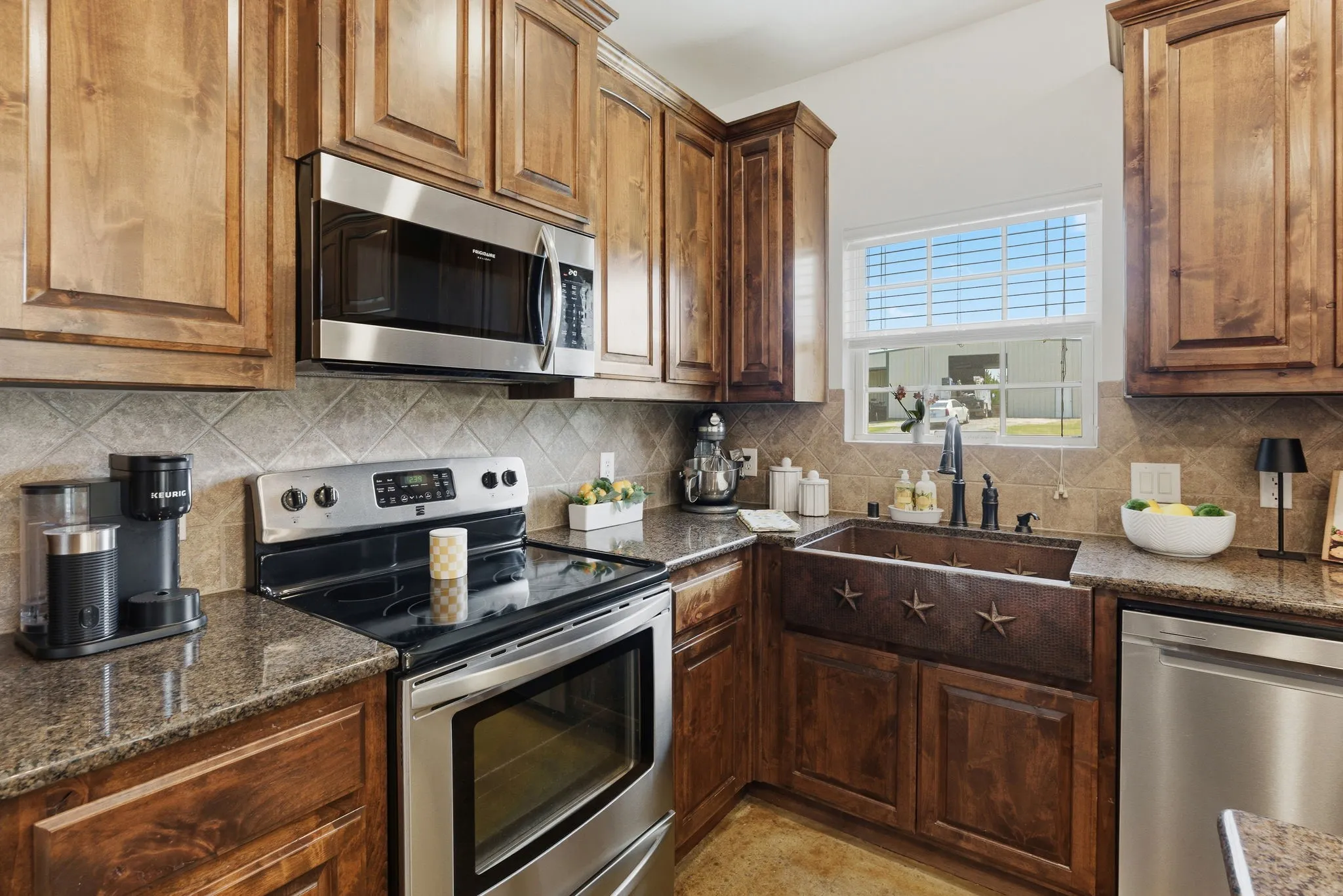 A distinctive apron-front sink with star detailing sits beneath a sunlit window, complemented by warm wood cabinetry, a diagonally laid tile backsplash, and conveniently arranged cooking and prep areas.